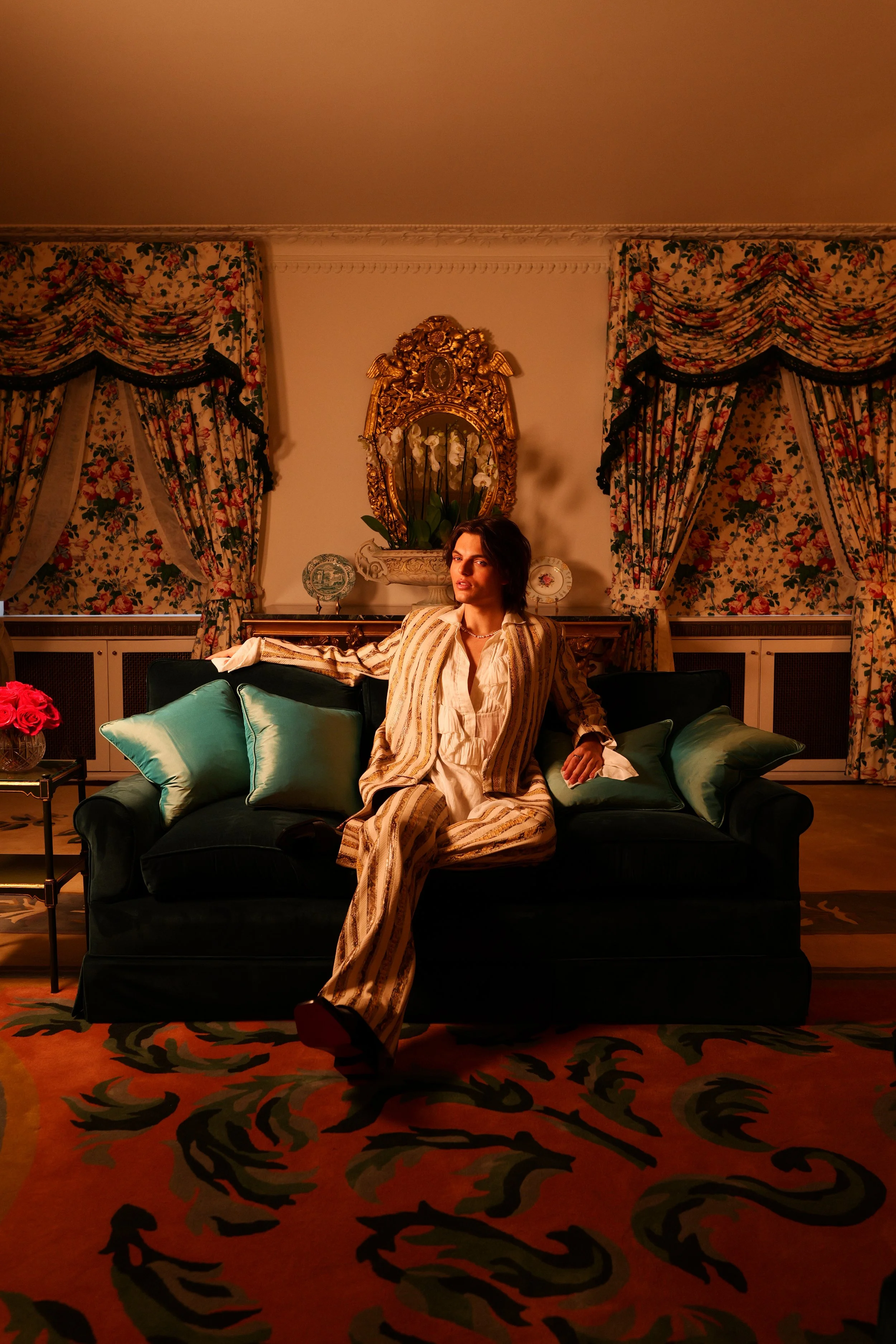 A young man with dark hair sitting on a black velvet sofa, wearing a light-colored, striped jacket and matching pants, in a room with floral curtains, a gold ornate mirror, and a rug with a red and black pattern.