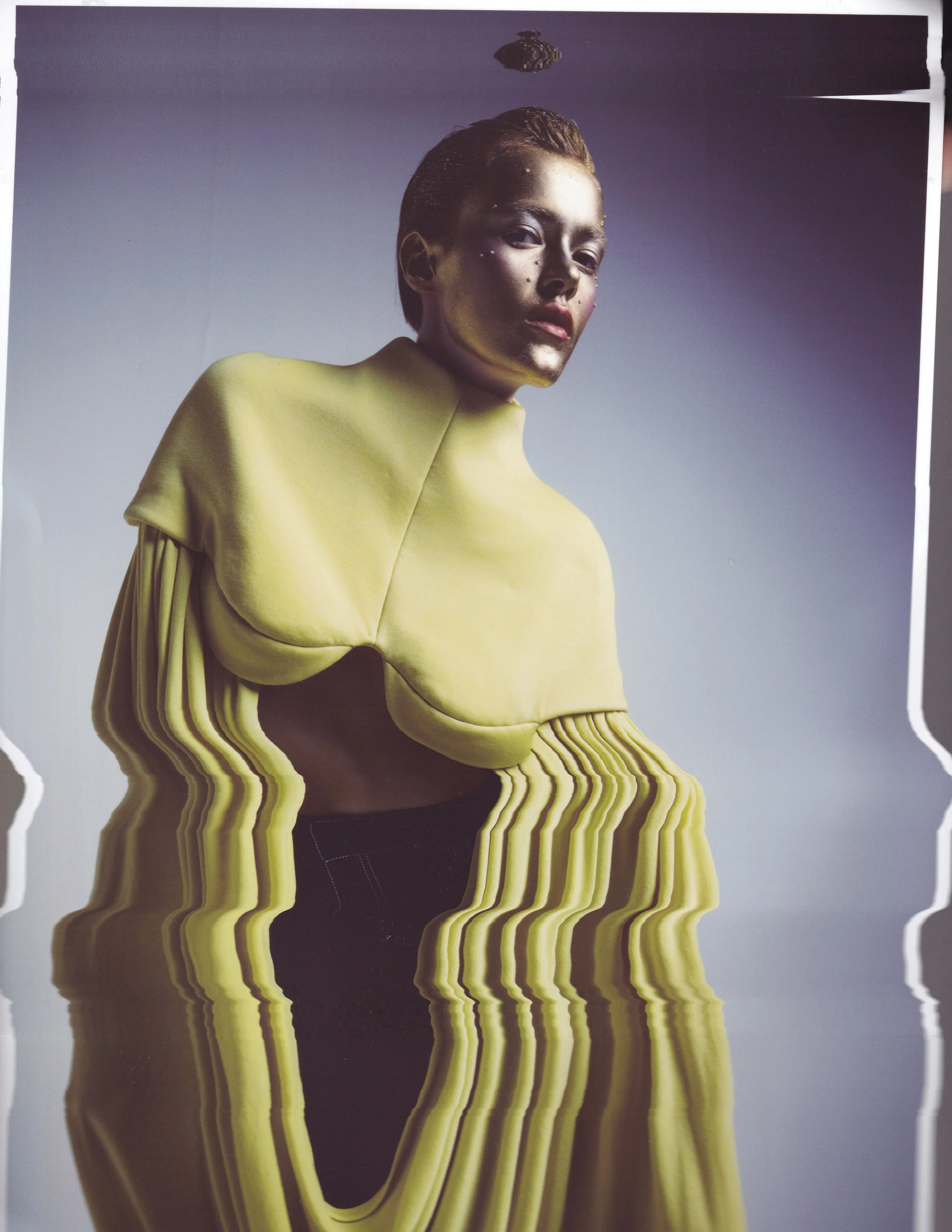 A woman with short brown hair and a serious expression wearing a yellow garment against a gradient blue background. The image has a warped, elongated, and distorted appearance.