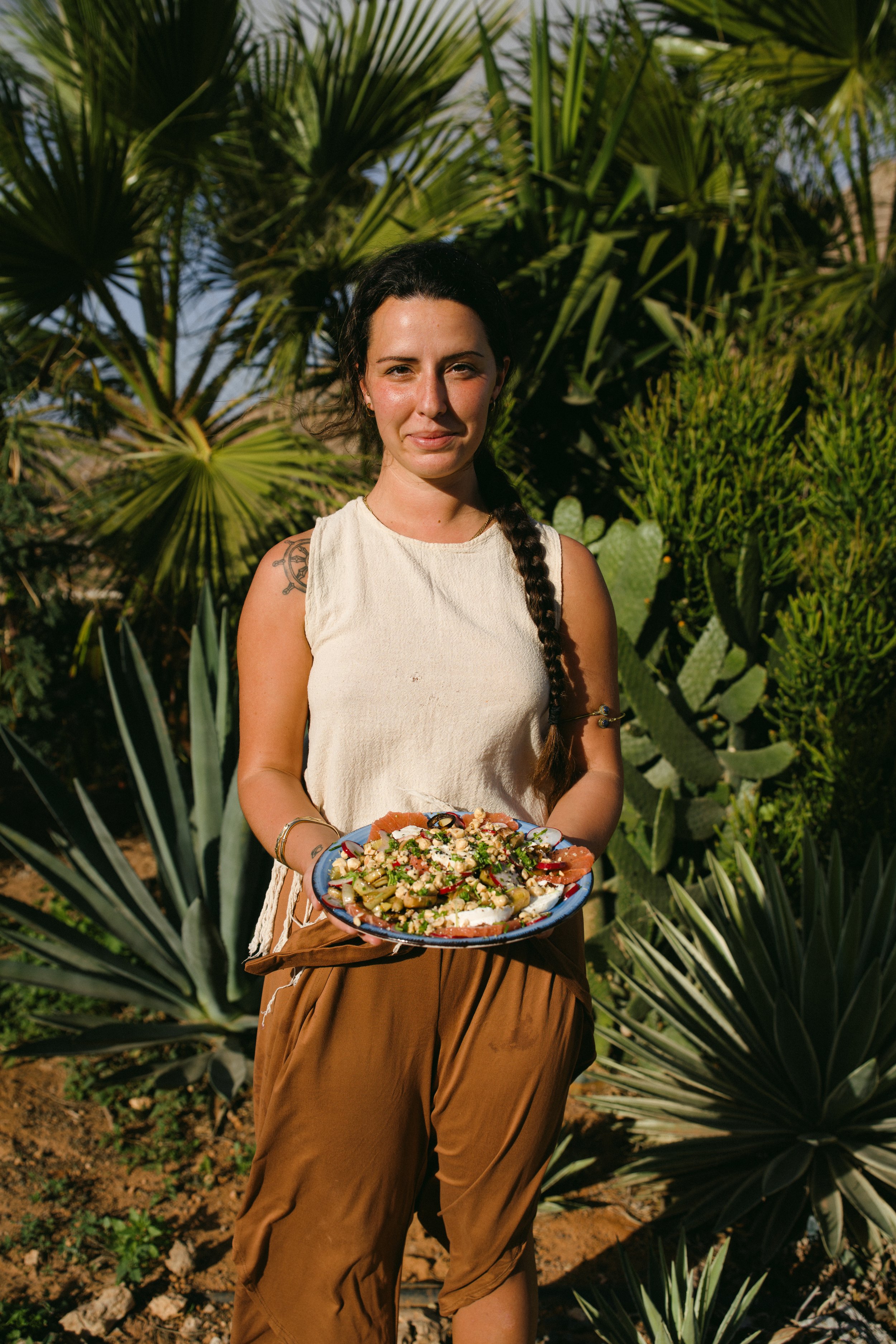 A young woman with long, curly brown hair holding a halved papaya on a beach at sunset, with the ocean and a clear sky in the background.