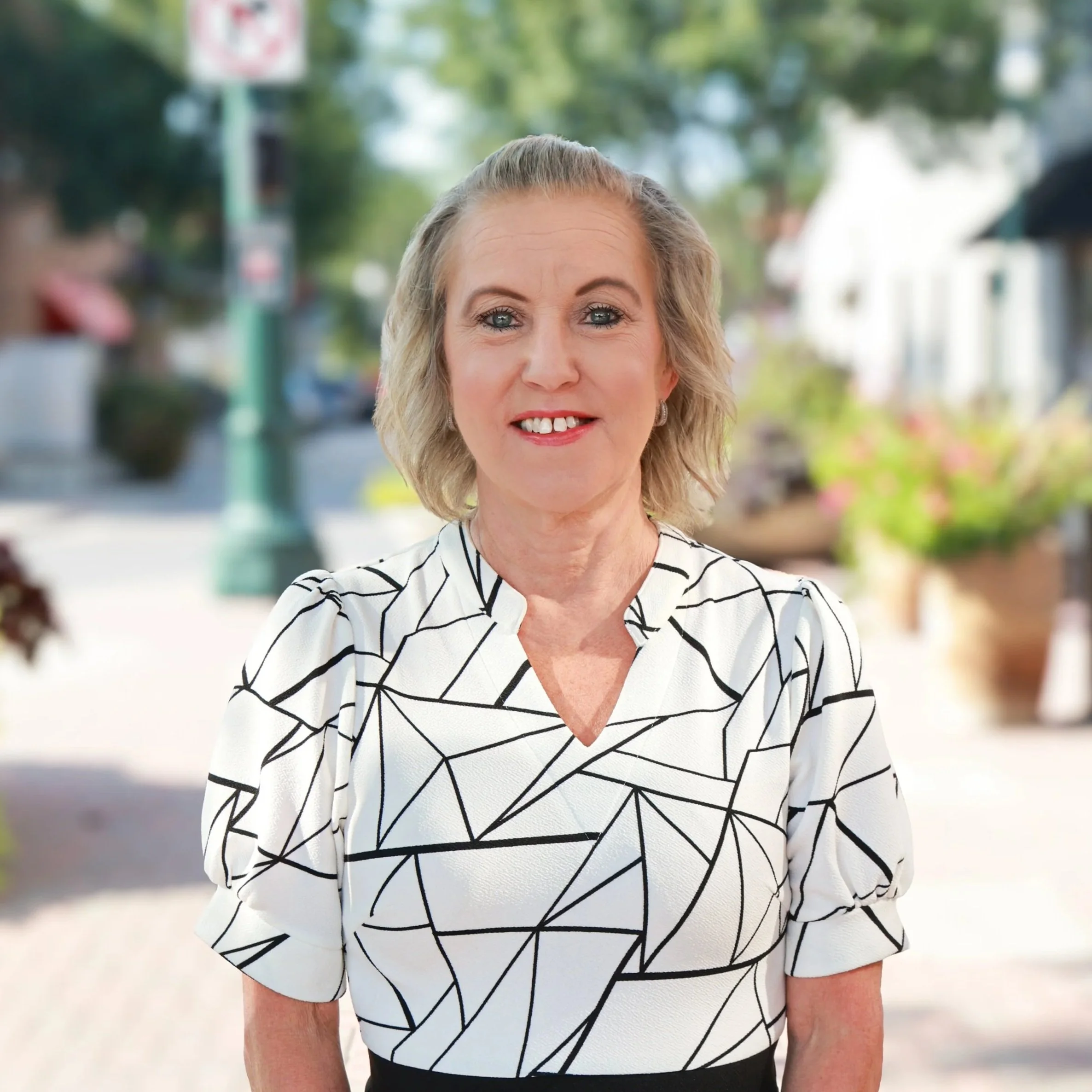 A woman with short blonde hair and blue eyes smiling, wearing a blue and black zebra-striped top, black necklace, earrings, and ring, standing in front of a stone wall.