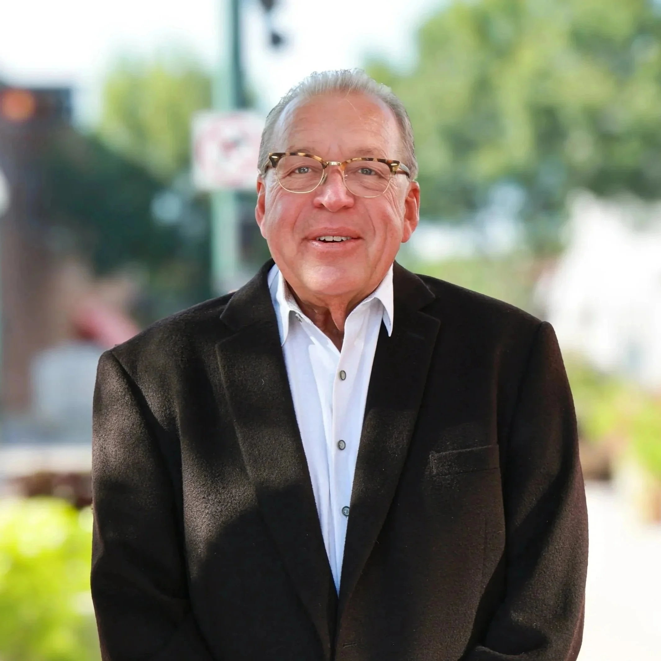 A smiling man with glasses in a black suit, white shirt, and patterned tie, standing against a plain gray background.