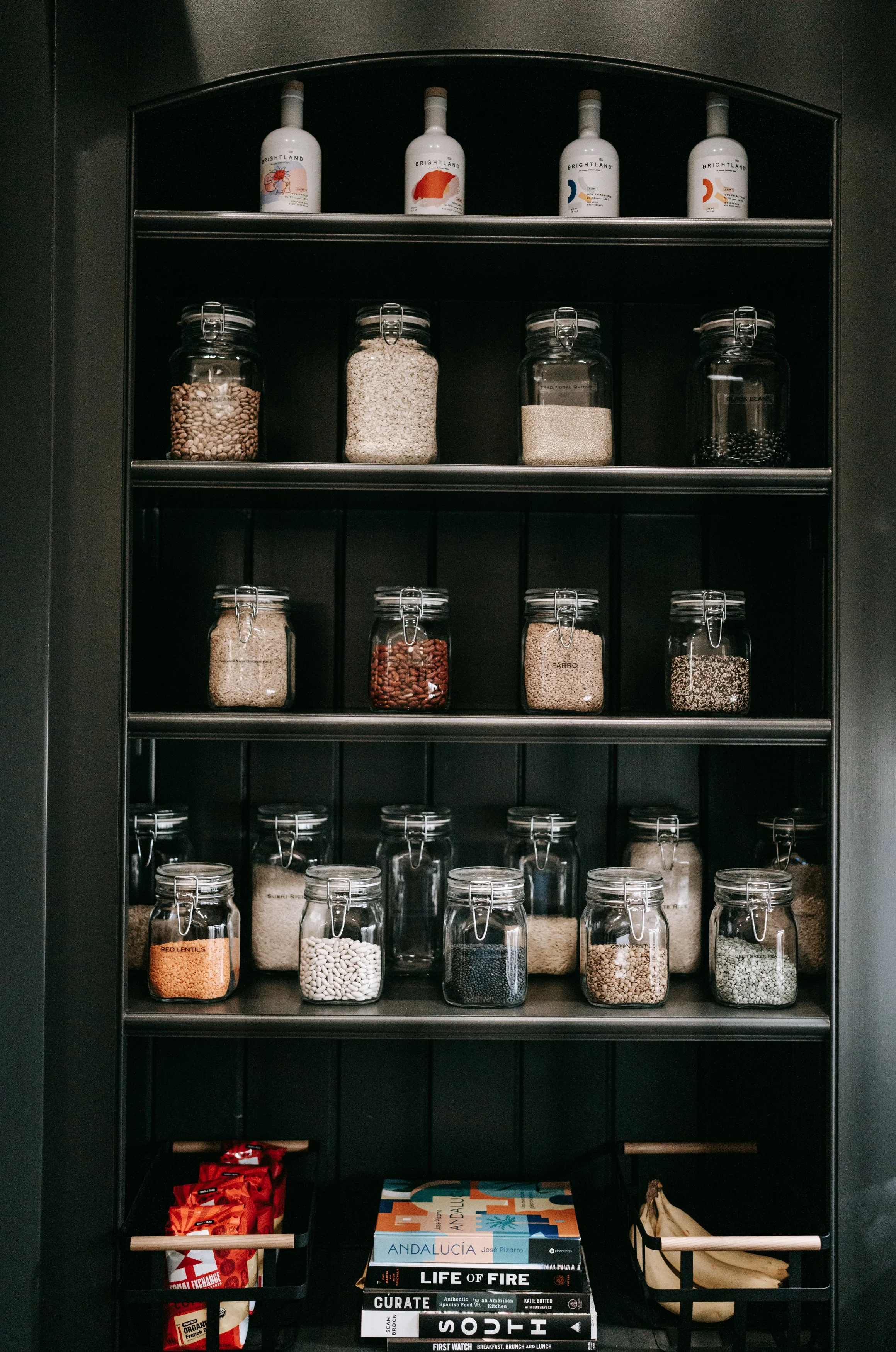 Pantry shelves with jars of grains and beans, bottles of olive oil on top, and baskets containing snacks and bananas at the bottom.