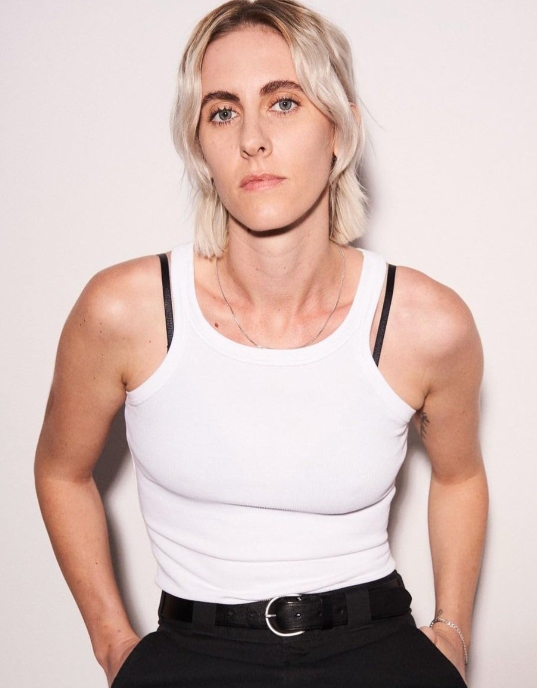 Person with short blonde hair wearing a white tank top and black pants, standing against a plain background.