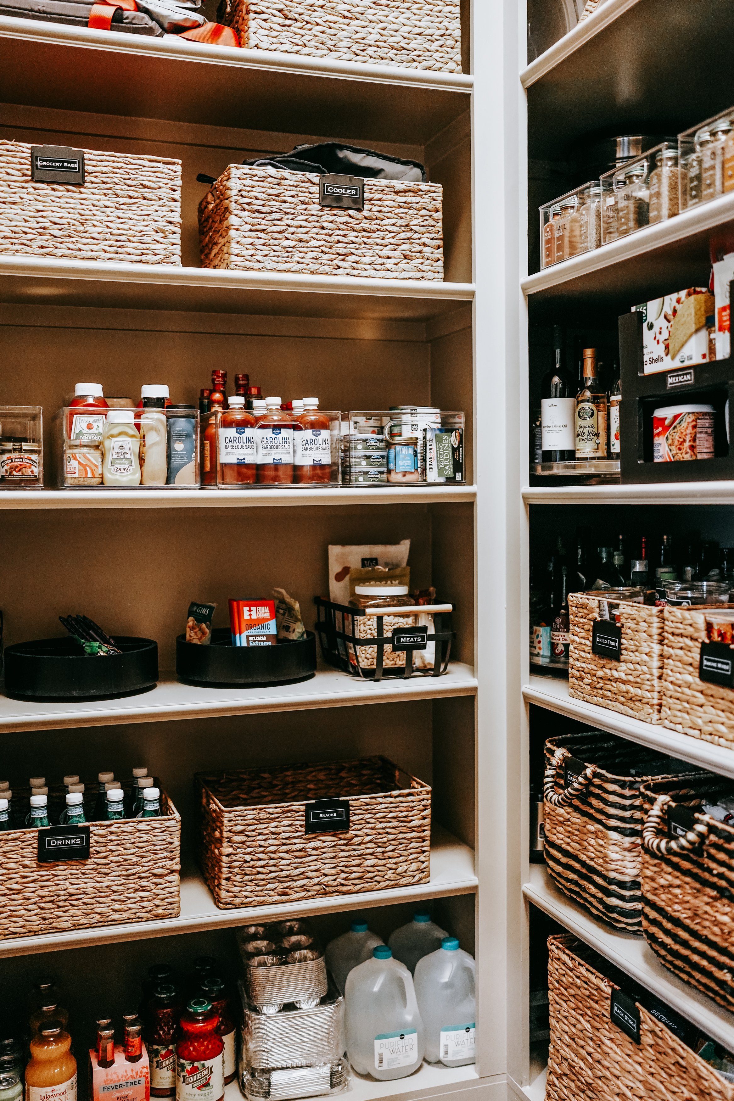 Organized pantry shelves with woven baskets, canned goods, condiments, sauces, water bottles, and labeled containers.