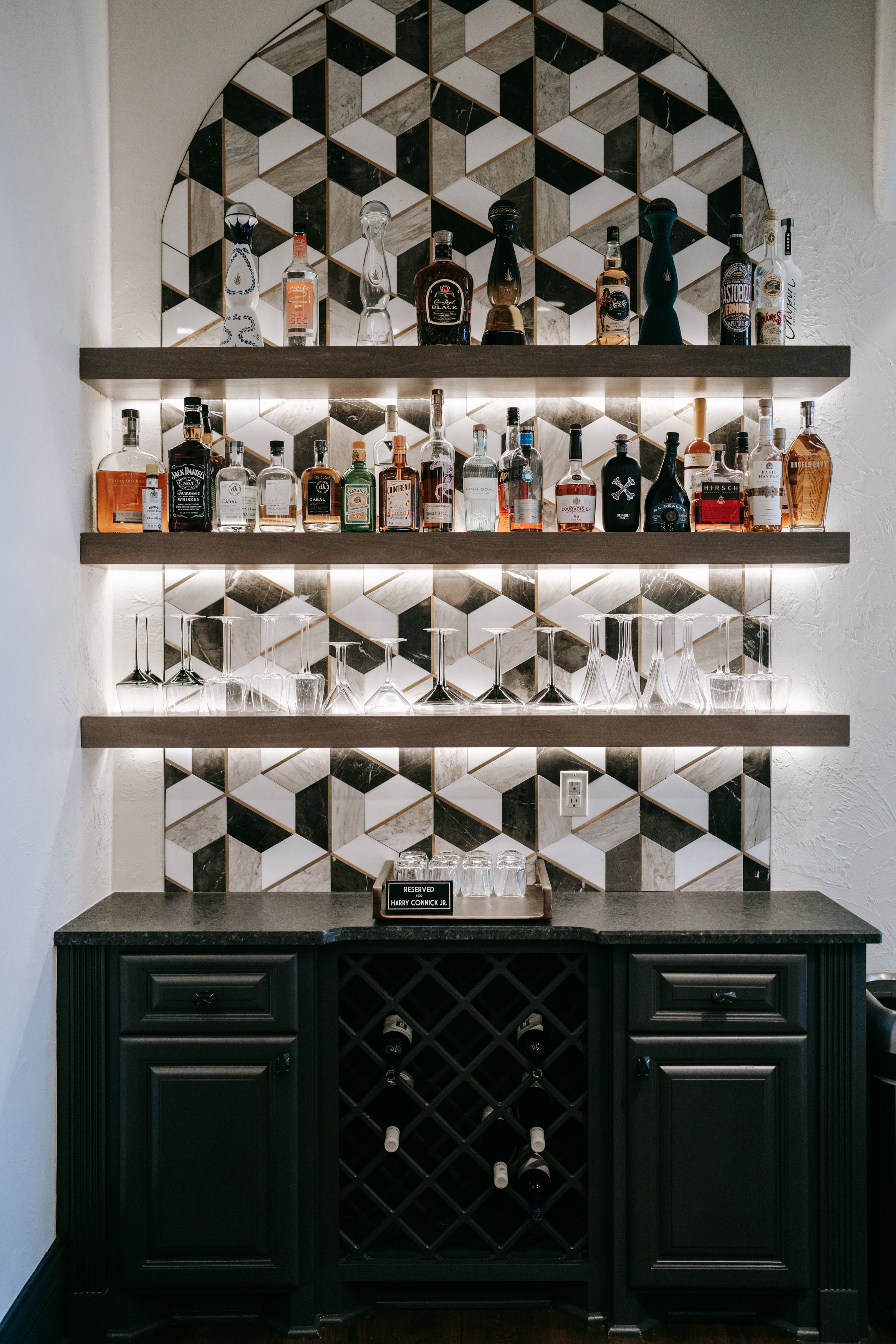 Home bar with shelves of liquor bottles and glasses, geometric tile backsplash, black cabinetry with wine rack and countertop, featuring a reserved sign.