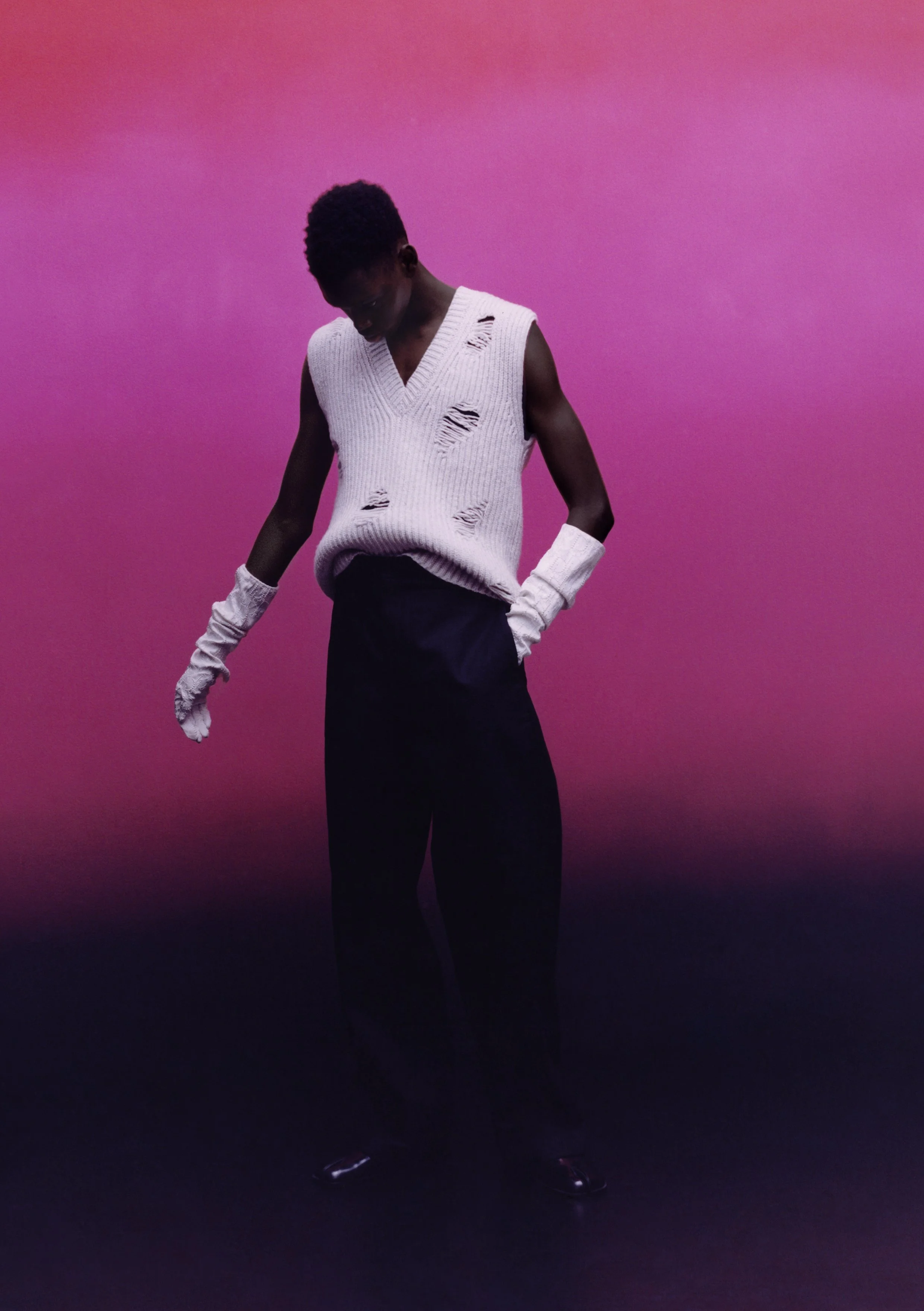 Person wearing a white, distressed sleeveless sweater, black pants, and gloves against a pink to black gradient background.