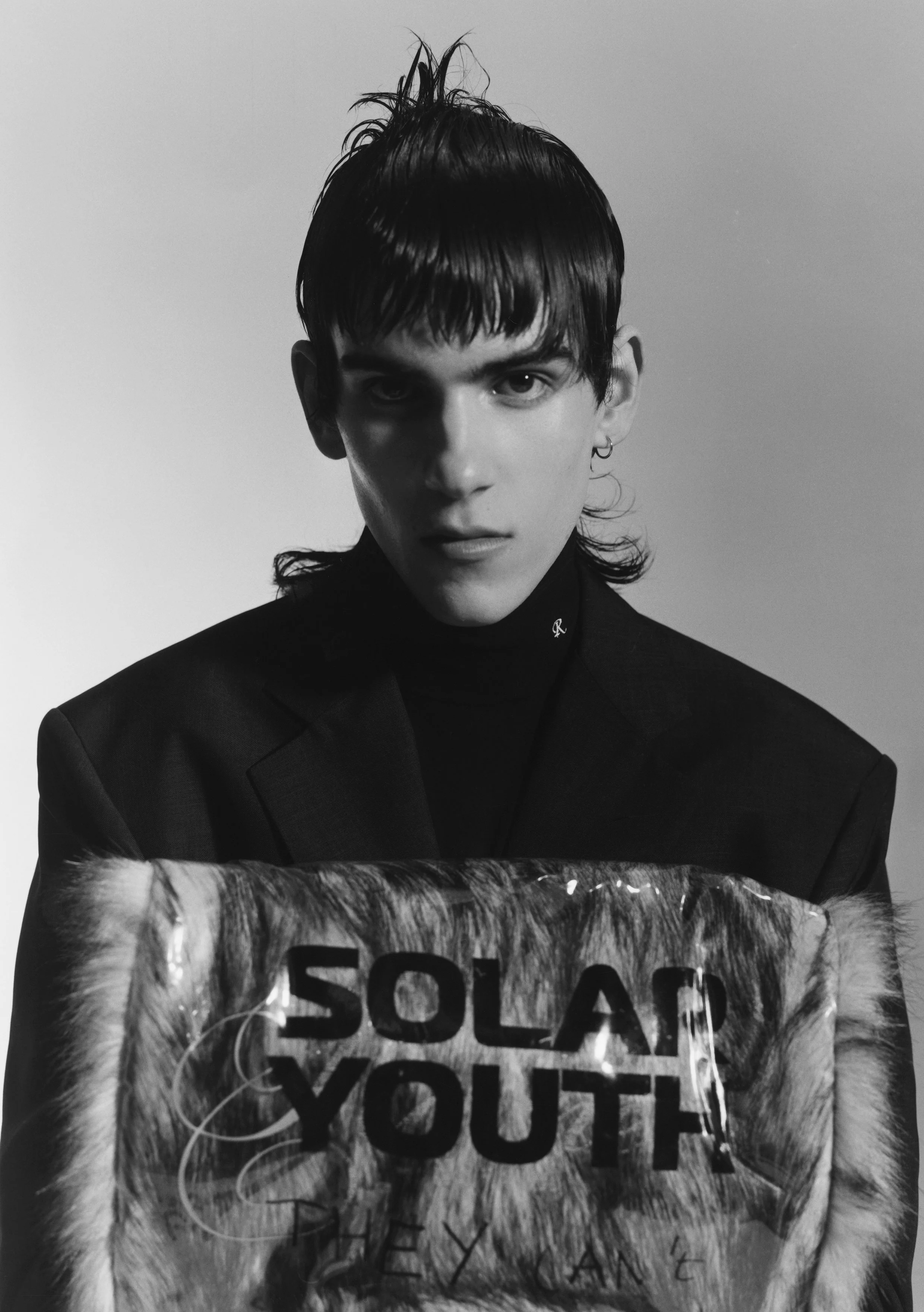 Black and white photo of a person with a fringe haircut, wearing a suit and holding a furry item with visible text "SOLAR YOUTH" and "THEY CAN'T."