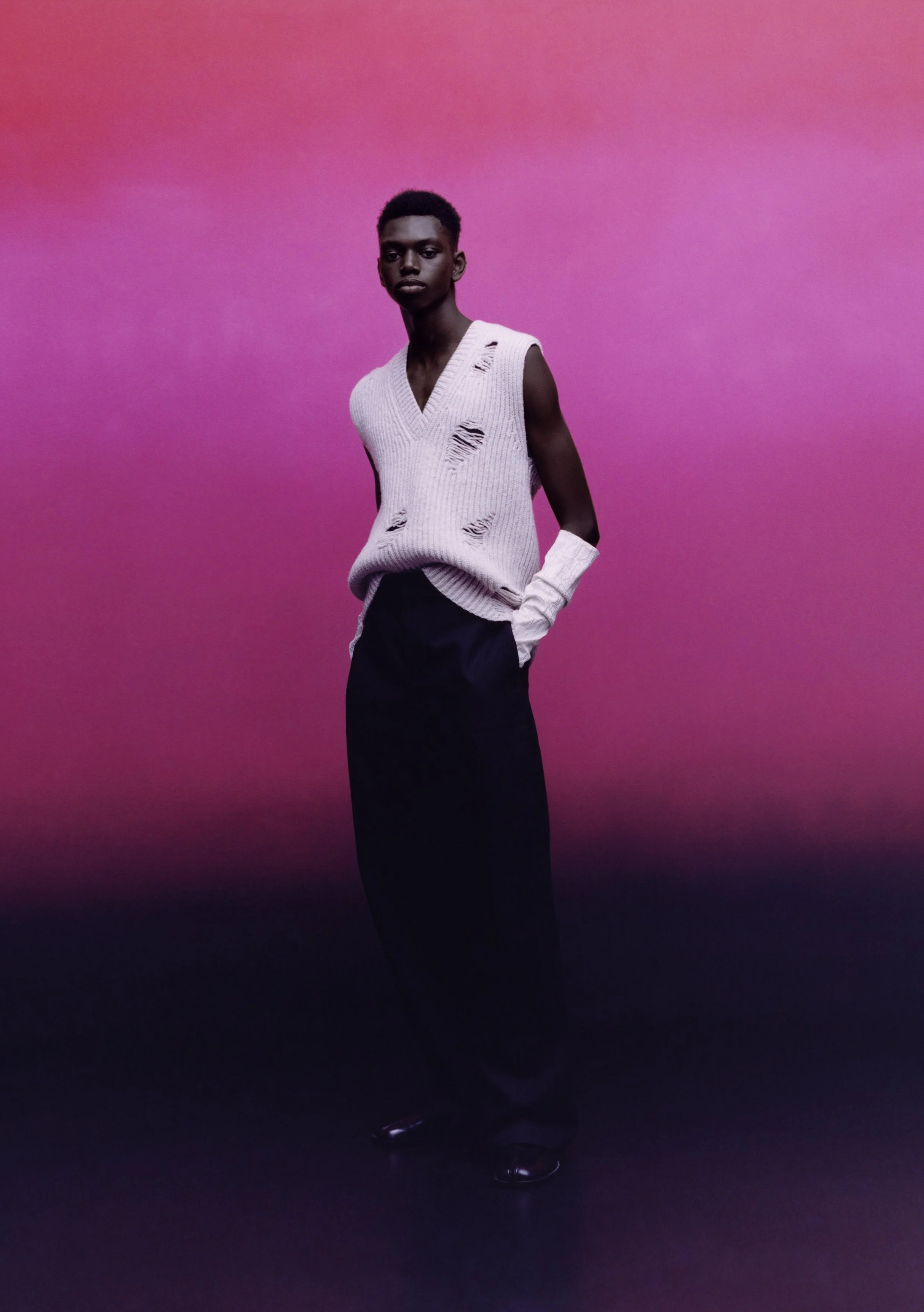 Person wearing a distressed sleeveless sweater and wide-leg pants, standing against a pink background.