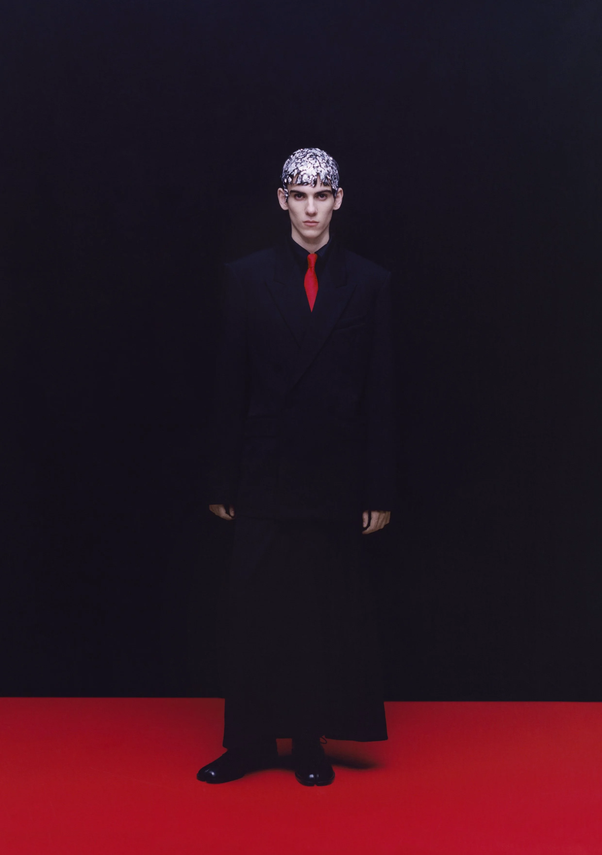 Person in black suit with red tie and patterned hat standing on red carpet against black background.