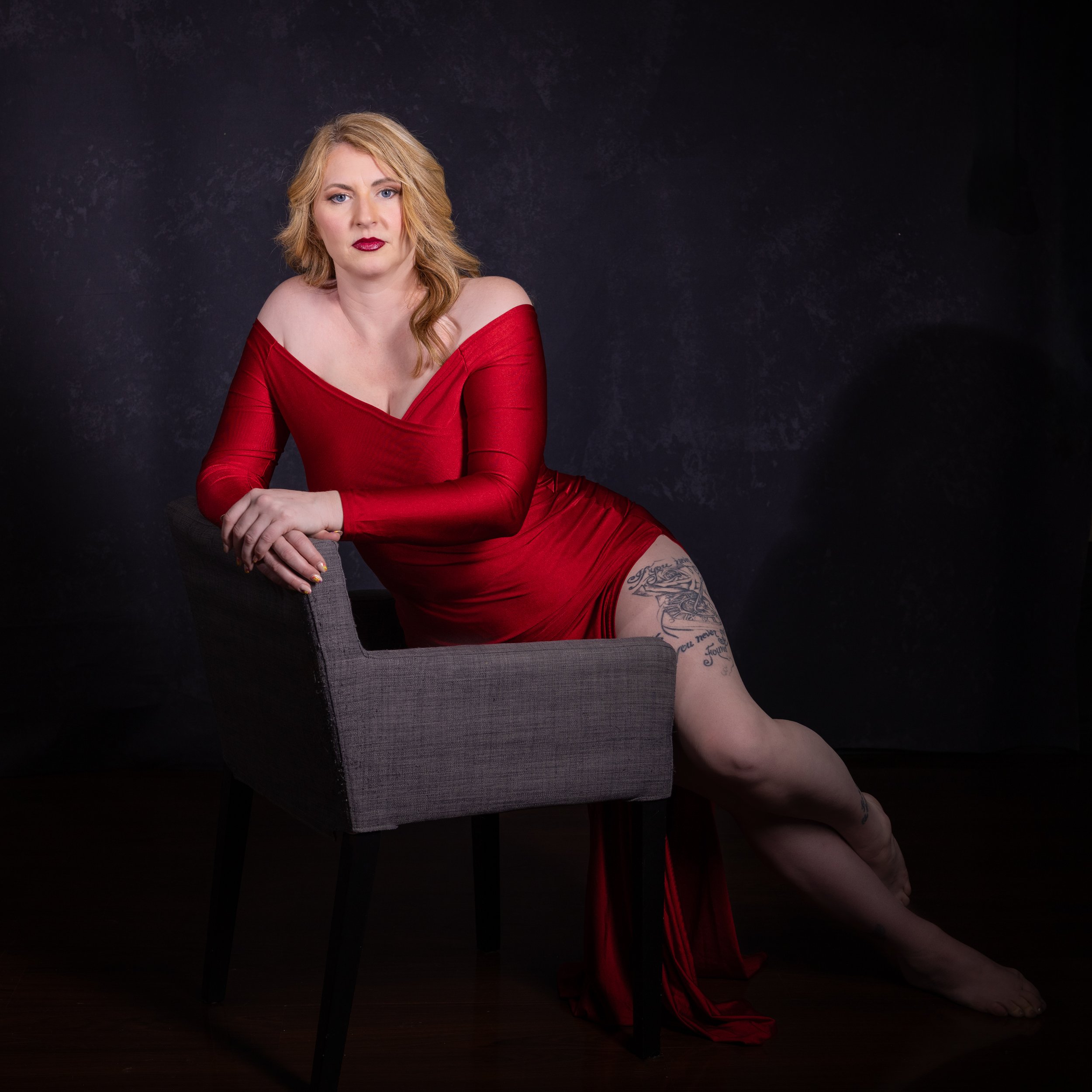 A woman with blonde hair and red lipstick wearing a fitted red dress sitting on a gray chair against a dark background.