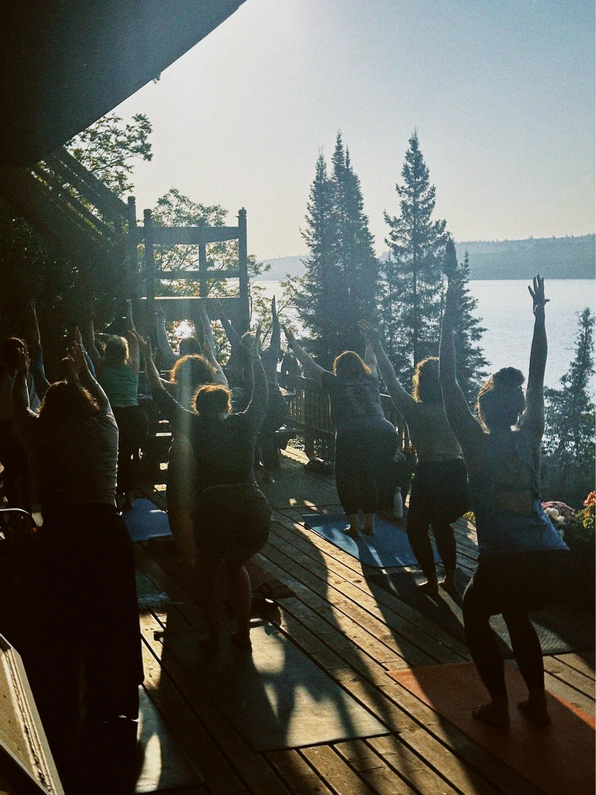 Group of people practicing yoga on a wooden deck by a lake, with trees and mountains in the background, during sunset.