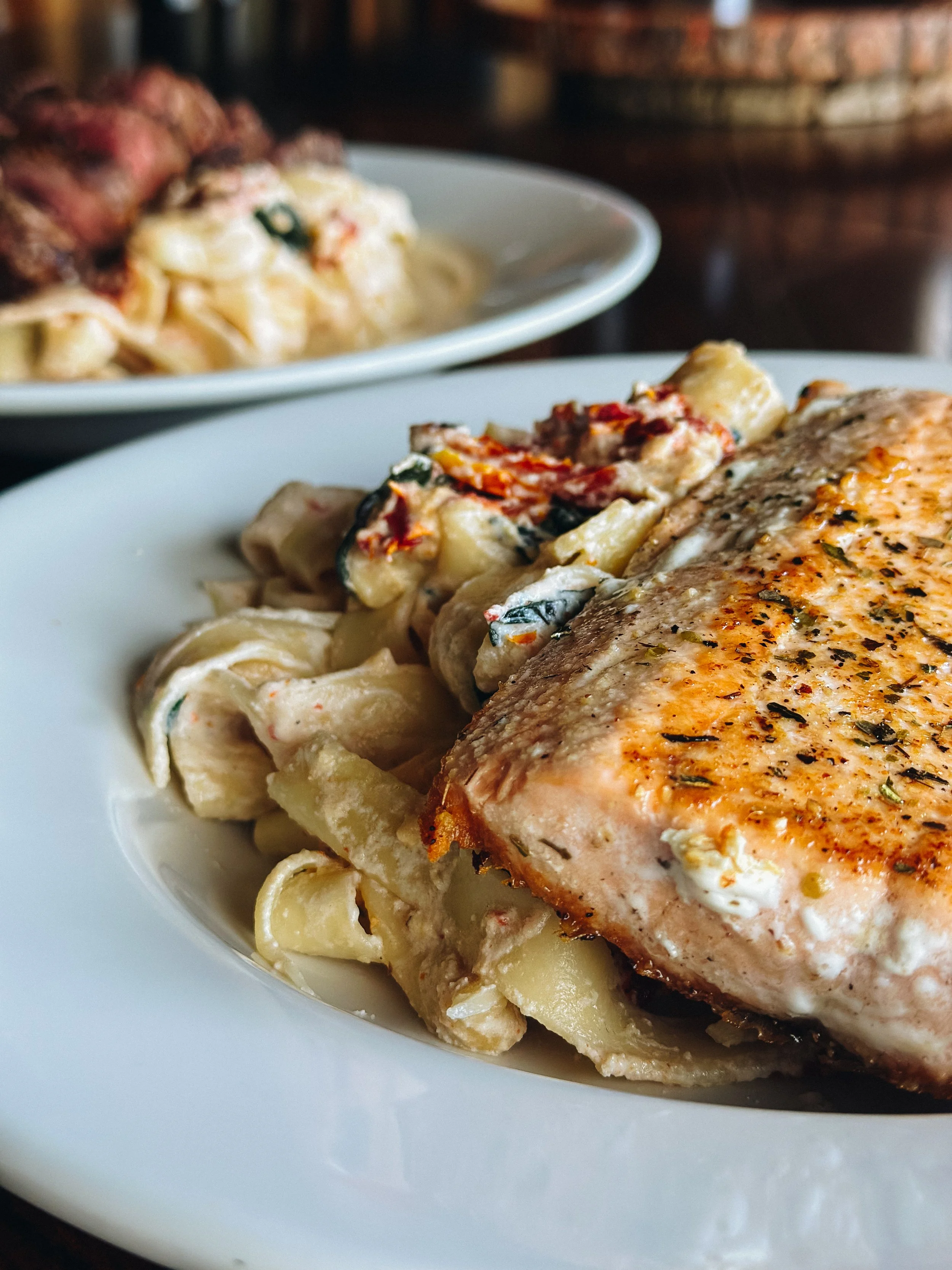 Close-up of a plate with grilled salmon, creamy pasta, and vegetable toppings, on a dark wooden table, with another plate of pasta in the background.