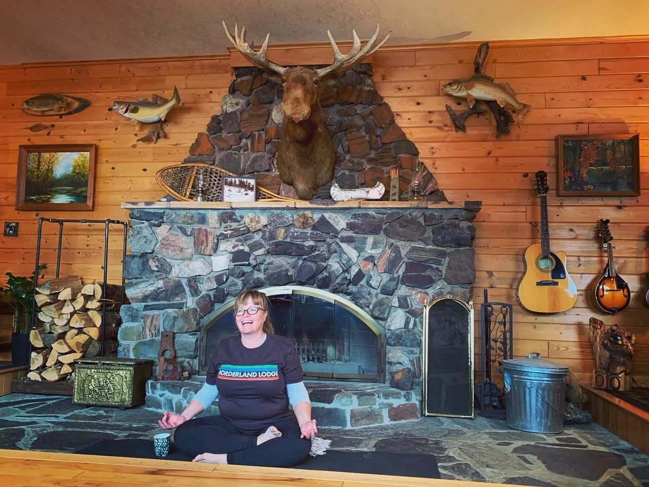 Woman sitting cross-legged and smiling in front of a rustic stone fireplace with a mounted moose head and surrounding wood-paneled walls decorated with fishing trophies, paintings, and guitars.
