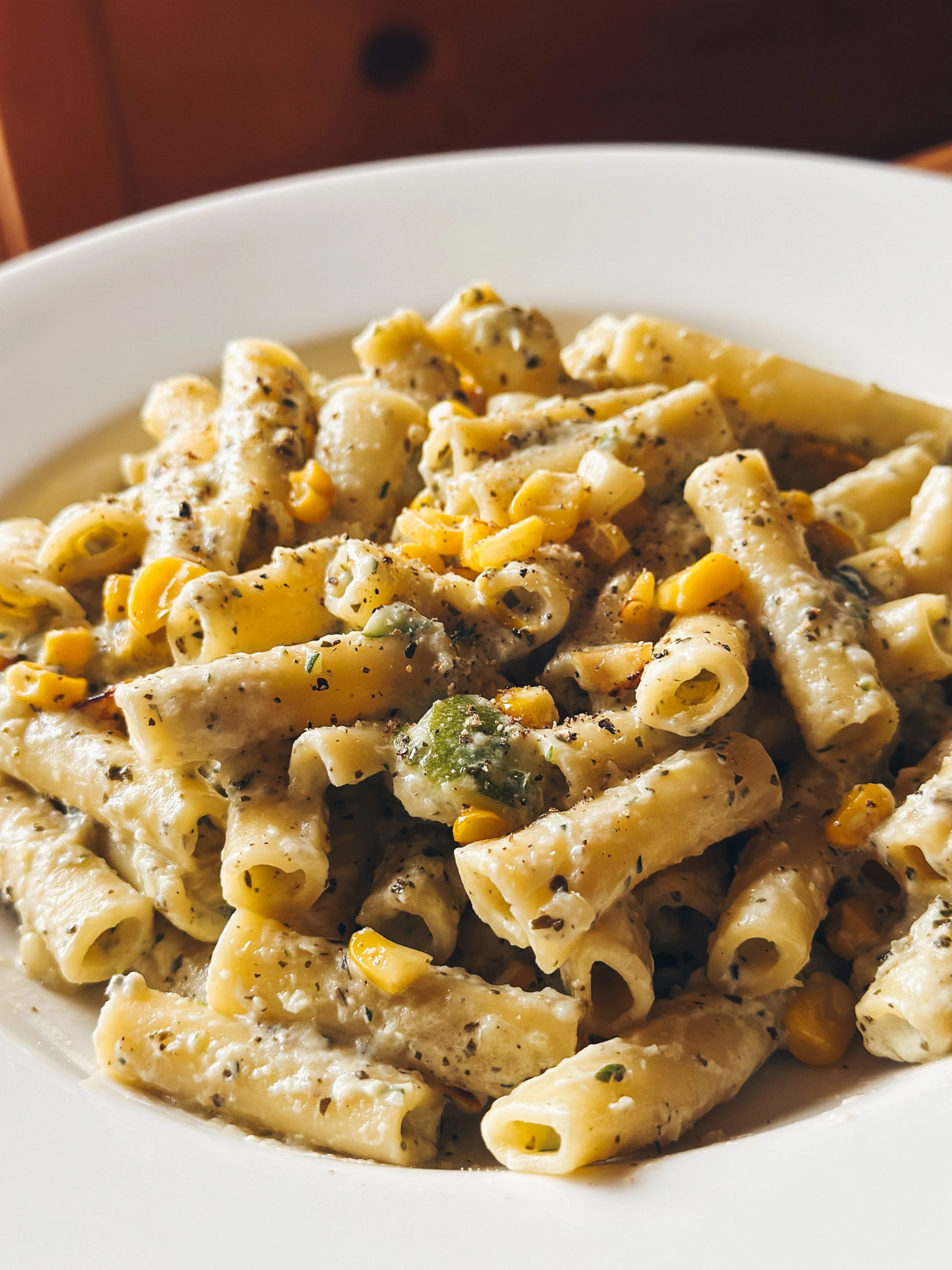 Creamy pasta with black pepper, corn, and green vegetables in a white bowl.