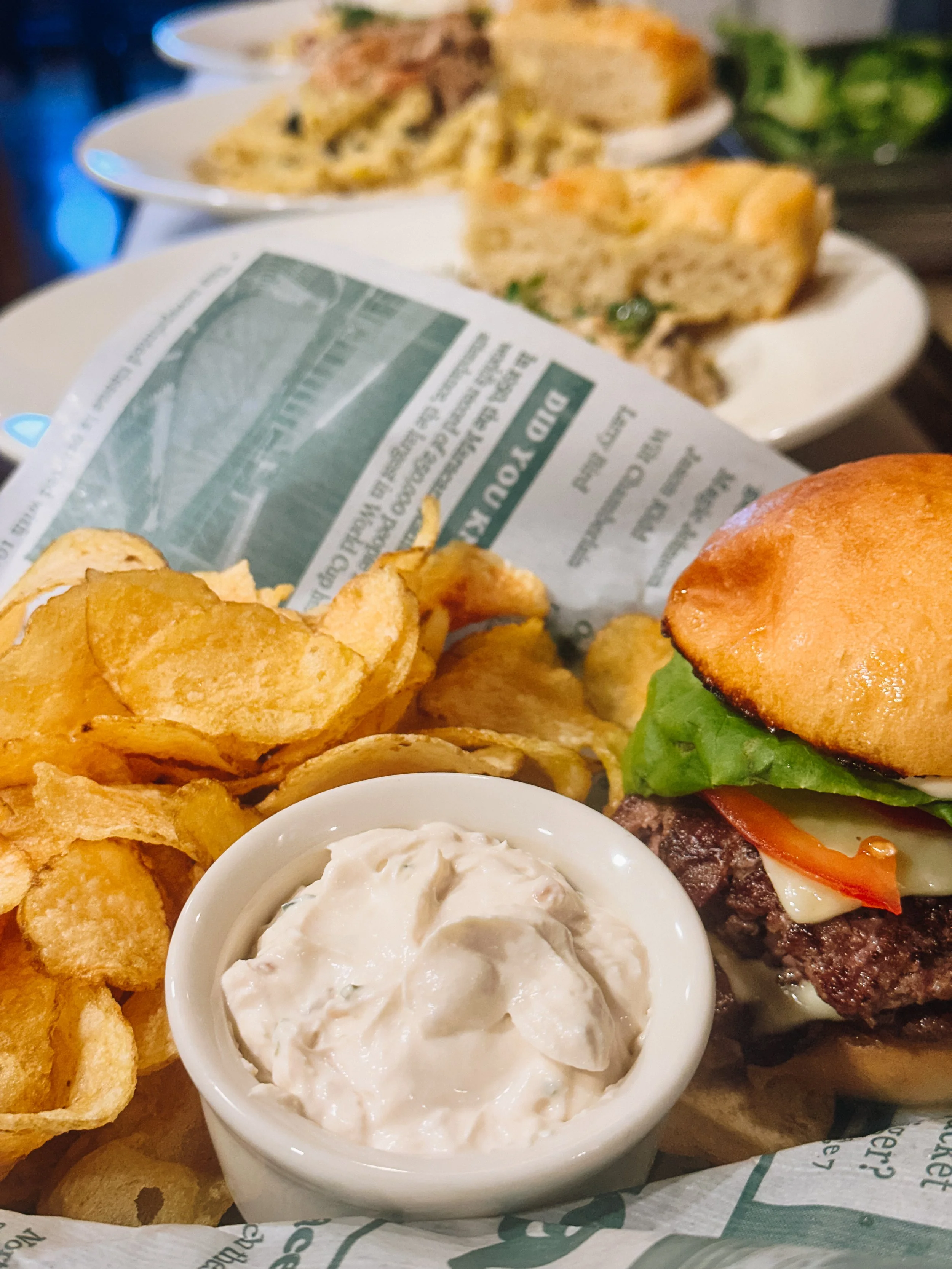 Close-up of a cheeseburger with lettuce, tomato, cheese, and a toasted bun, served with potato chips and a side of creamy dip. In the background, there are plates of pasta and a slice of quiche on a table.