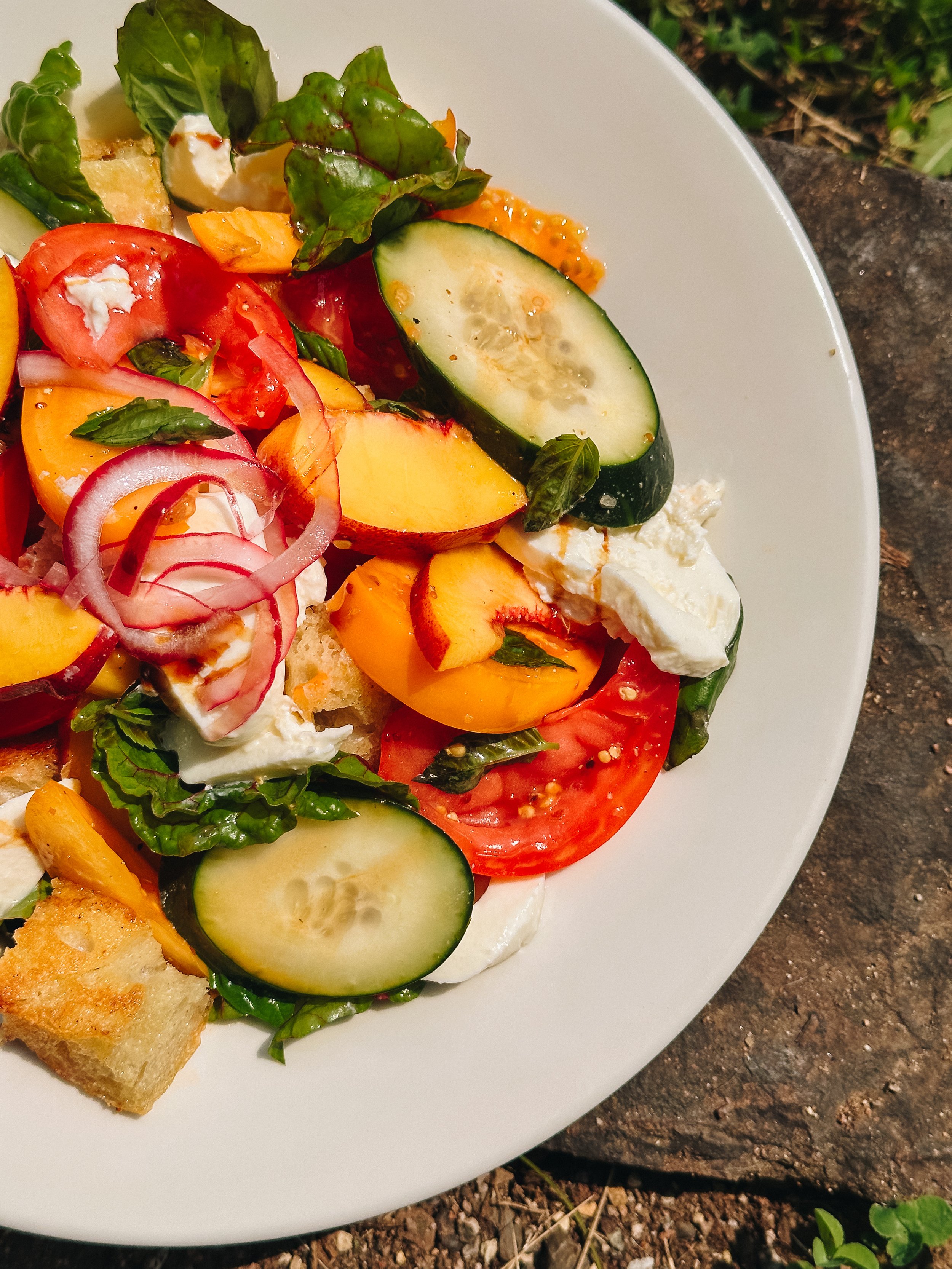 Close-up of a fresh vegetable salad on a white plate with sliced cucumbers, tomatoes, red onions, bell peppers, leafy greens, croutons, and dollops of feta cheese.