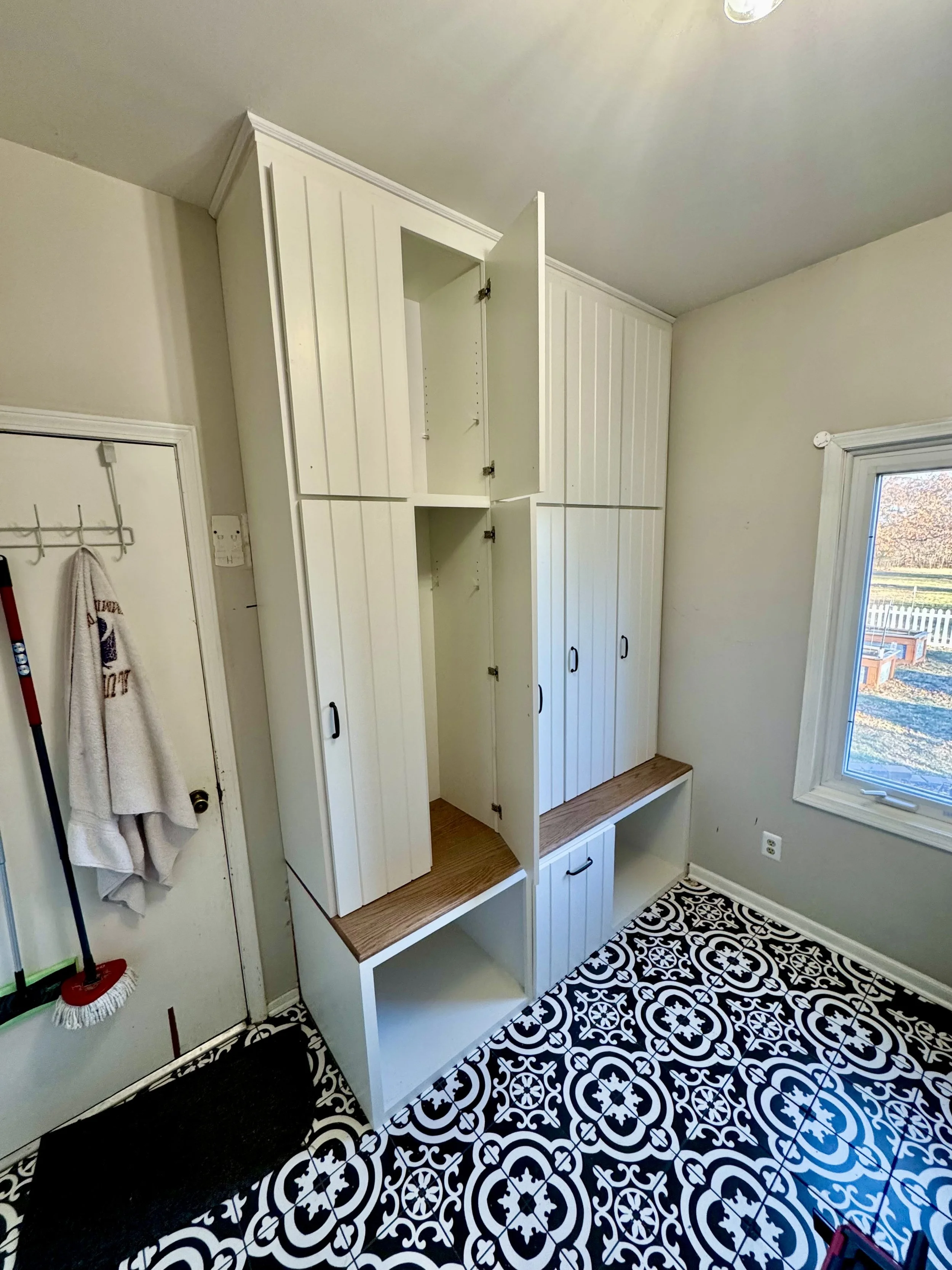 White built-in storage cabinets and bench in a kitchen or mudroom with black and white patterned tile floor and a window