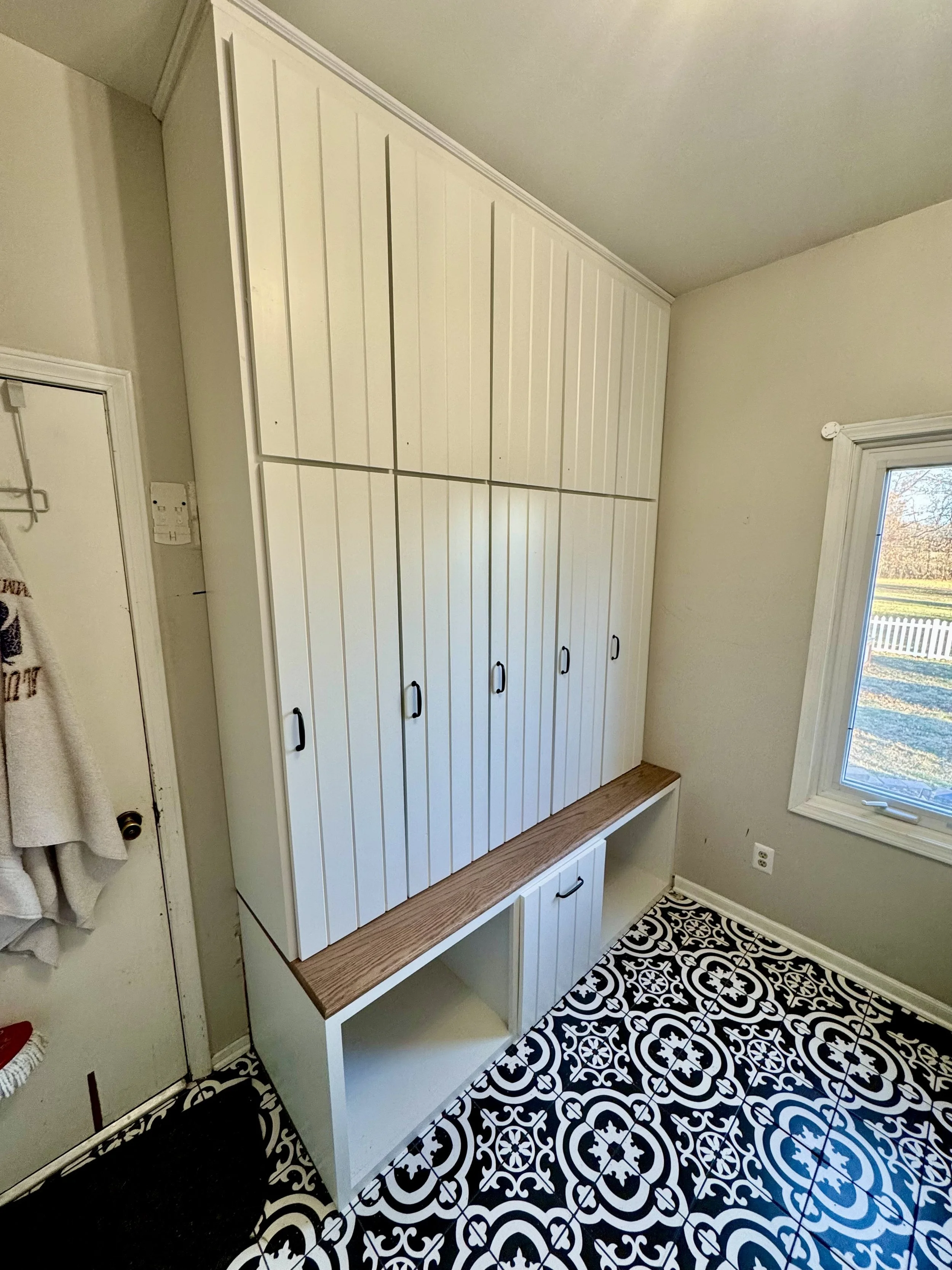 White cabinets with black handles above and below a wooden countertop in a room with black and white patterned tile floor and a window.