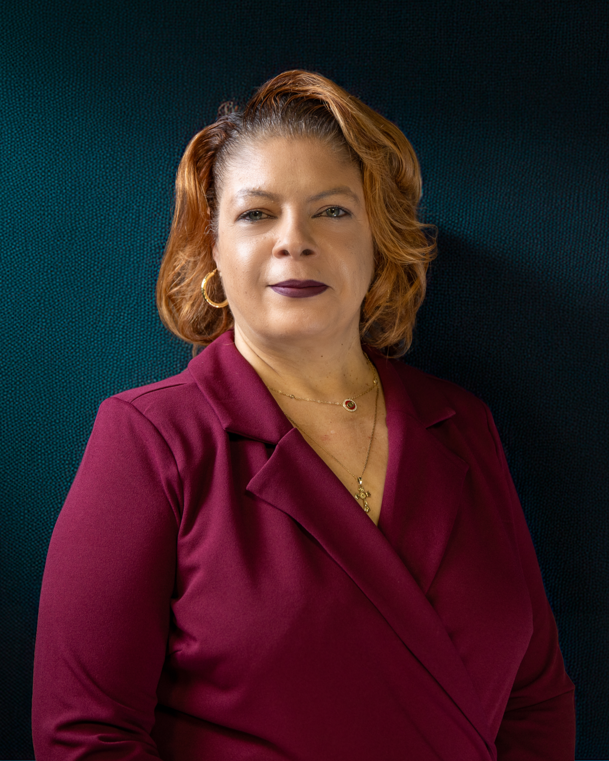 A woman with red, curly hair in a professional portrait, wearing a black blazer over a zebra-patterned blouse, gold hoop earrings, and a gold necklace with a cross pendant.