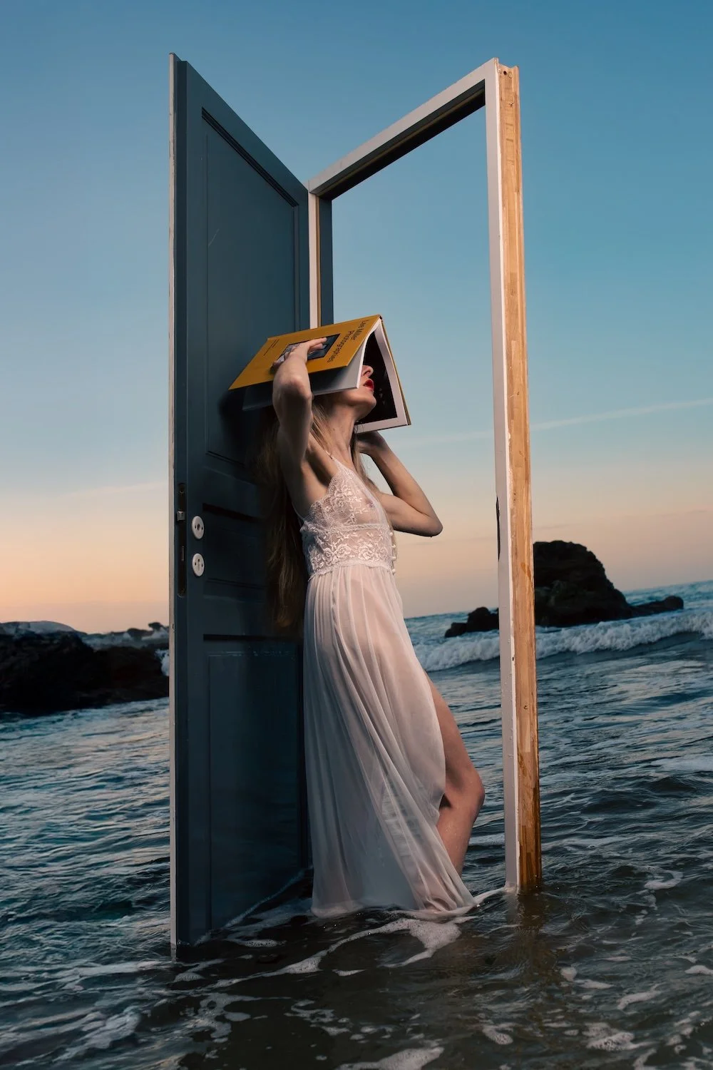 Femme en nuisette blanche debout dans l'océan, appuyée contre une porte ouverte avec un livre ouvert sur le visage, photographie poétique et surréaliste.
