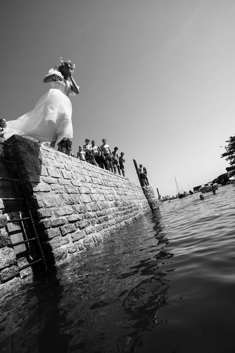 Mariée en robe blanche sautant depuis la jetée du port dans la mer lors d'un reportage photo de mariage documentaire.