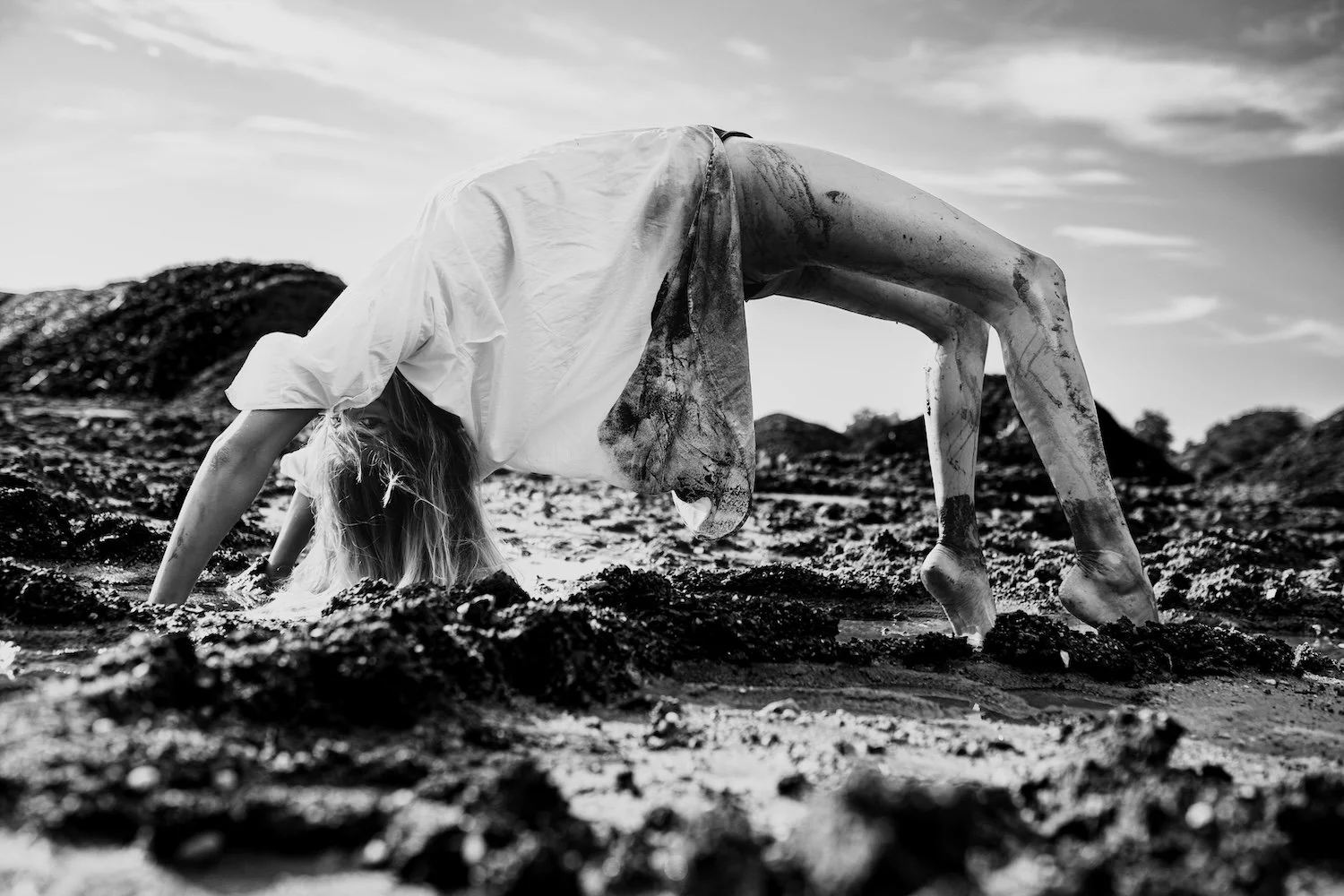 Photographie noir et blanc artistique en contre-plongée, montrant une femme en posture de pont inversée, mains et pieds nus dans la boue texturée d'un chantier à Nantes, portant une chemise blanche tachetée de boue.