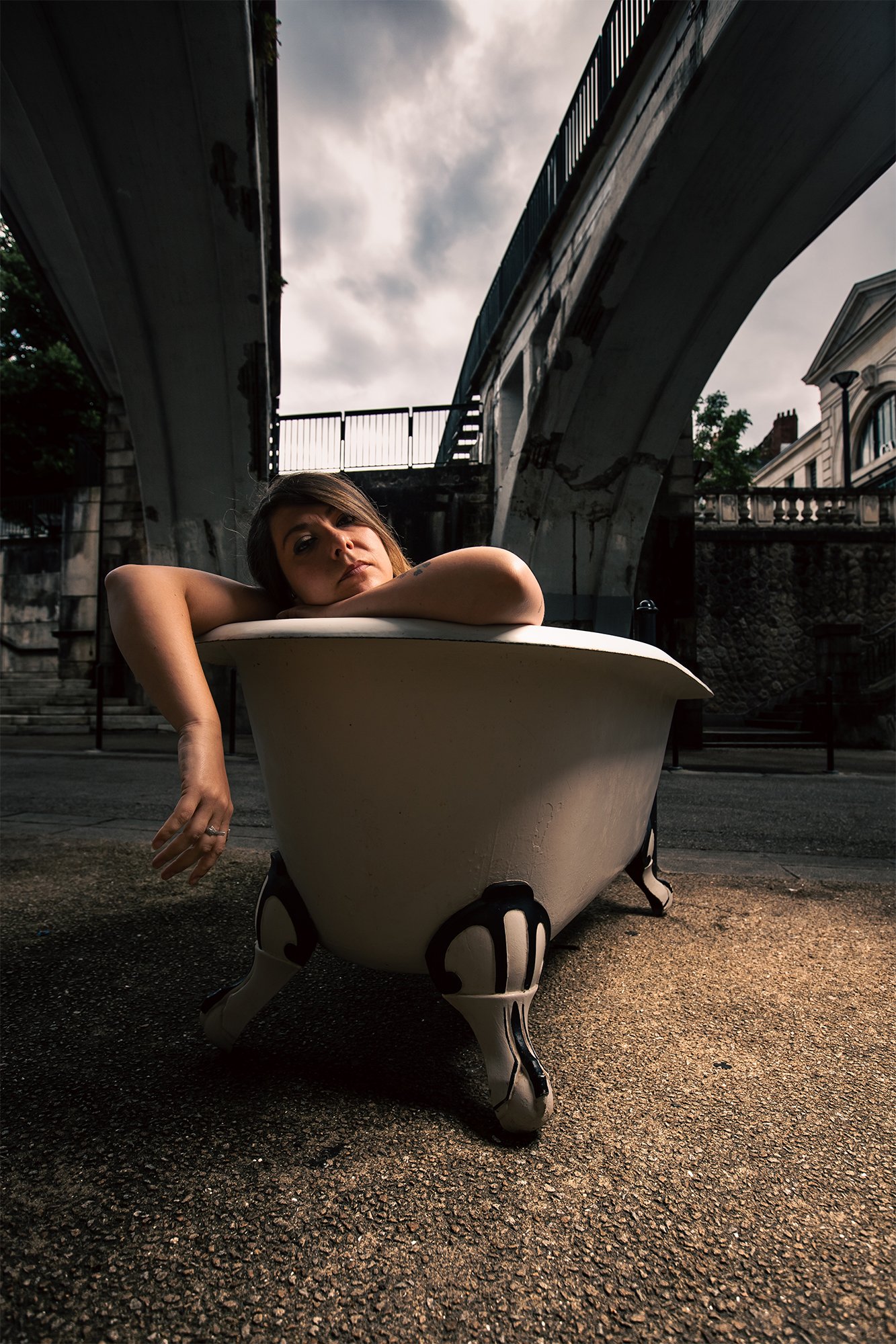 Photographie conceptuelle d’une femme allongée dans une baignoire en extérieur sous un pont urbain, explorant le contraste entre confort et environnement brut