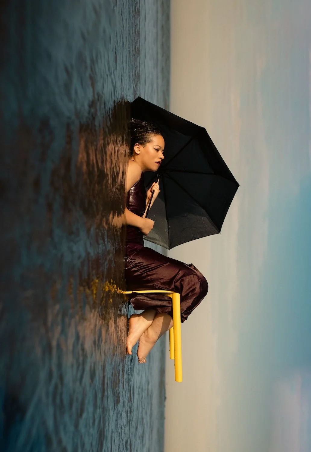 Portrait artistique surréaliste d'une femme assise sur un tabouret jaune, tenant un parapluie noir, flottant entre une surface d'eau et un ciel abstrait, photographie conceptuelle à Nantes.