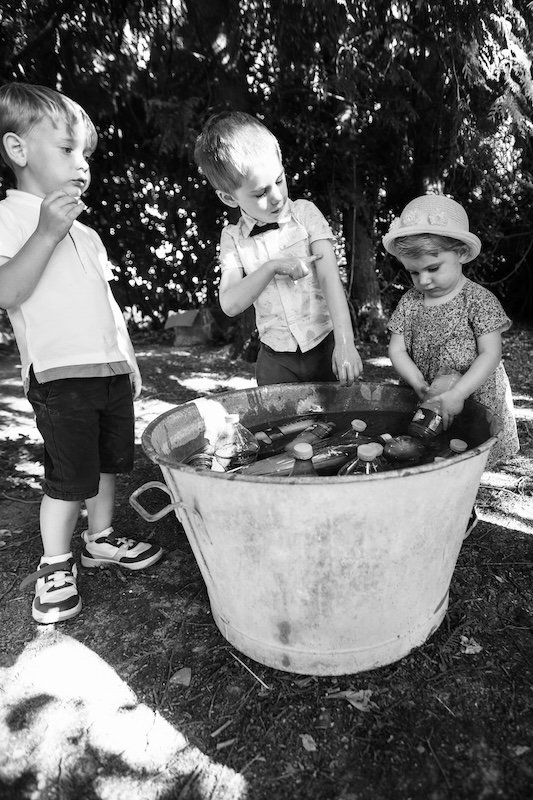 Trois enfants en tenue de fête s'amusant autour d'une vieille bassine en zinc remplie de bouteilles d'eau rafraîchissantes lors d'une chaude journée de mariage, photographié en noir et blanc par François Mougel.