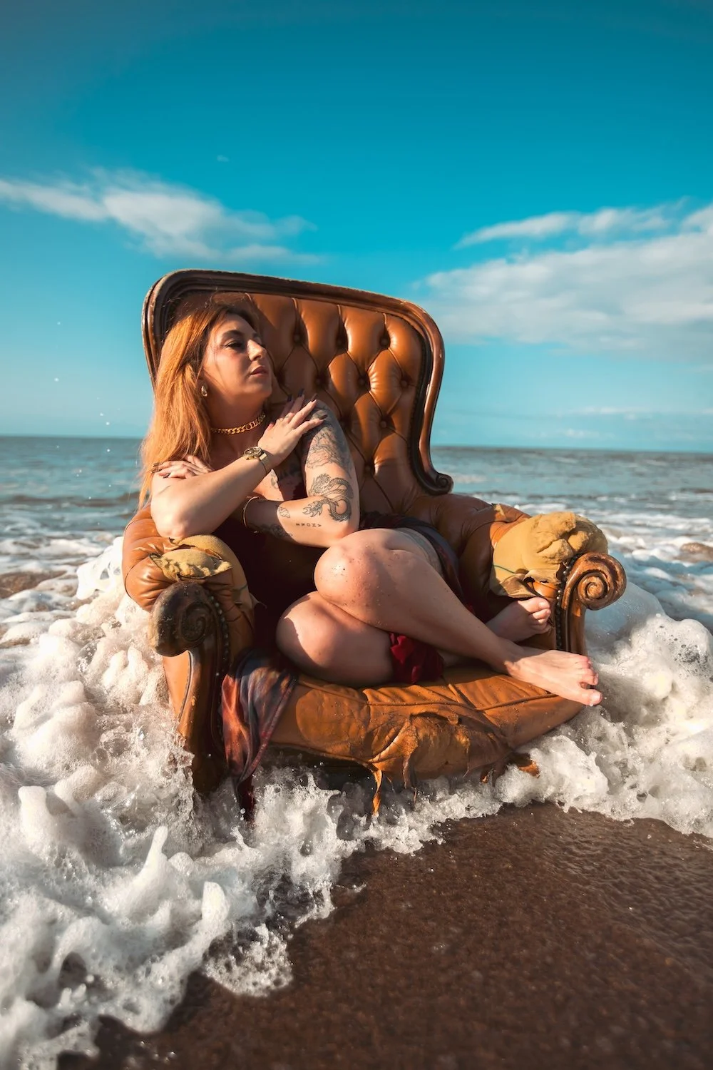Femme tatouée assise dans un fauteuil en cuir capitonné posé au bord du rivage, pieds nus entourés par l'écume des vagues sur la plage.