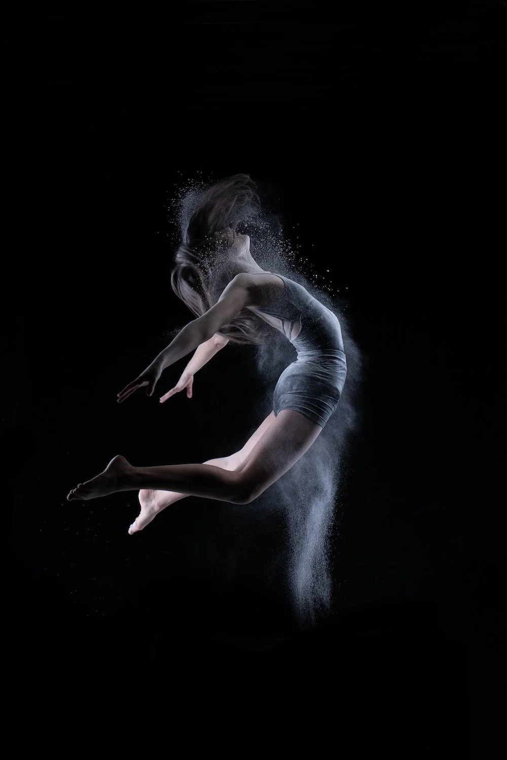 Danseuse en body gris capturée en plein saut dynamique sur fond noir, entourée d'un nuage de poudre blanche en mouvement.