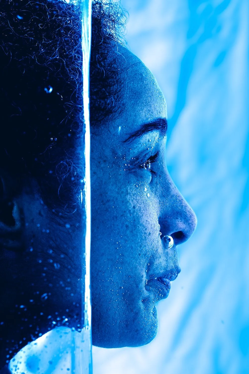 Profil artistique en contre-plongée et éclairage bleu saturé d'une personne aux cheveux bouclés, dont le visage est traversé par un filet d'eau et parsemé de bulles, photographie conceptuelle à Nantes.