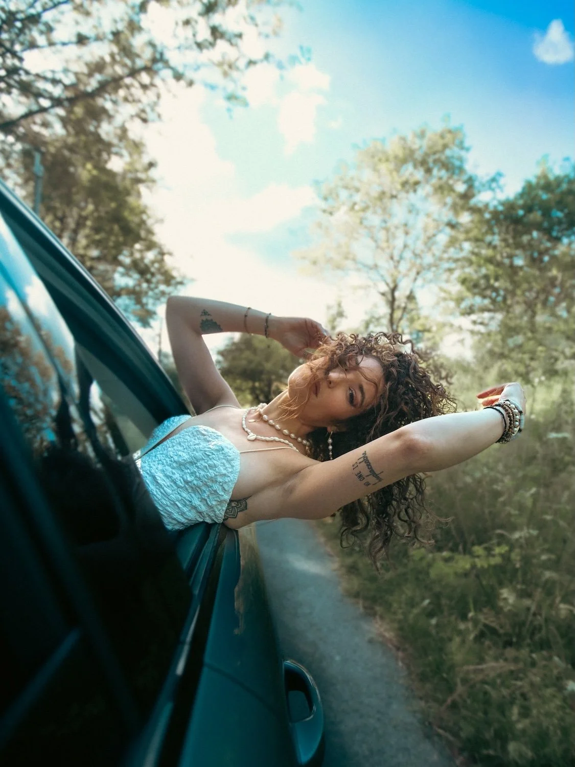 Jeune femme penchée à la fenêtre d'une voiture, lascive, avec les bras levés, regardant vers l'objectif, portant des bijoux et des tatouages, sous un ciel bleu avec des arbres en arrière-plan.