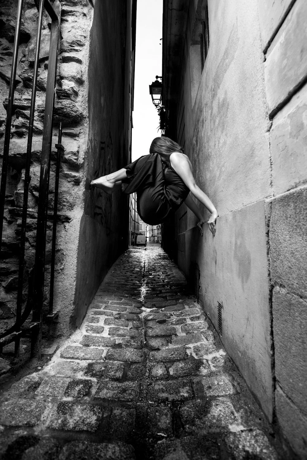 Femme suspendue en équilibre acrobatique entre deux murs d'une ruelle pavée à Nantes, photographie noir et blanc.