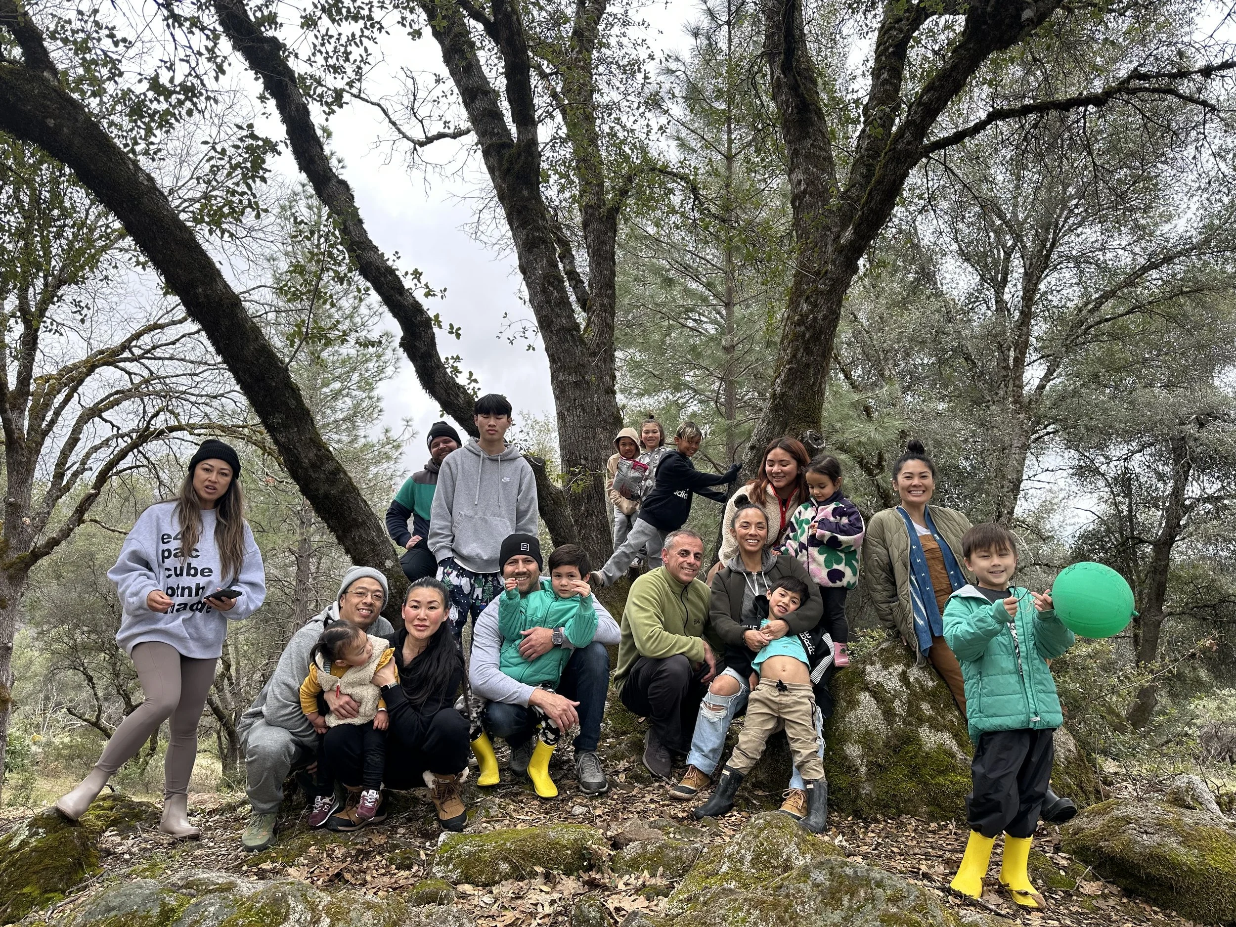 Group of people, including children and adults, gathered outdoors on rocks and tree roots in a forested area, smiling and posing for a photo.