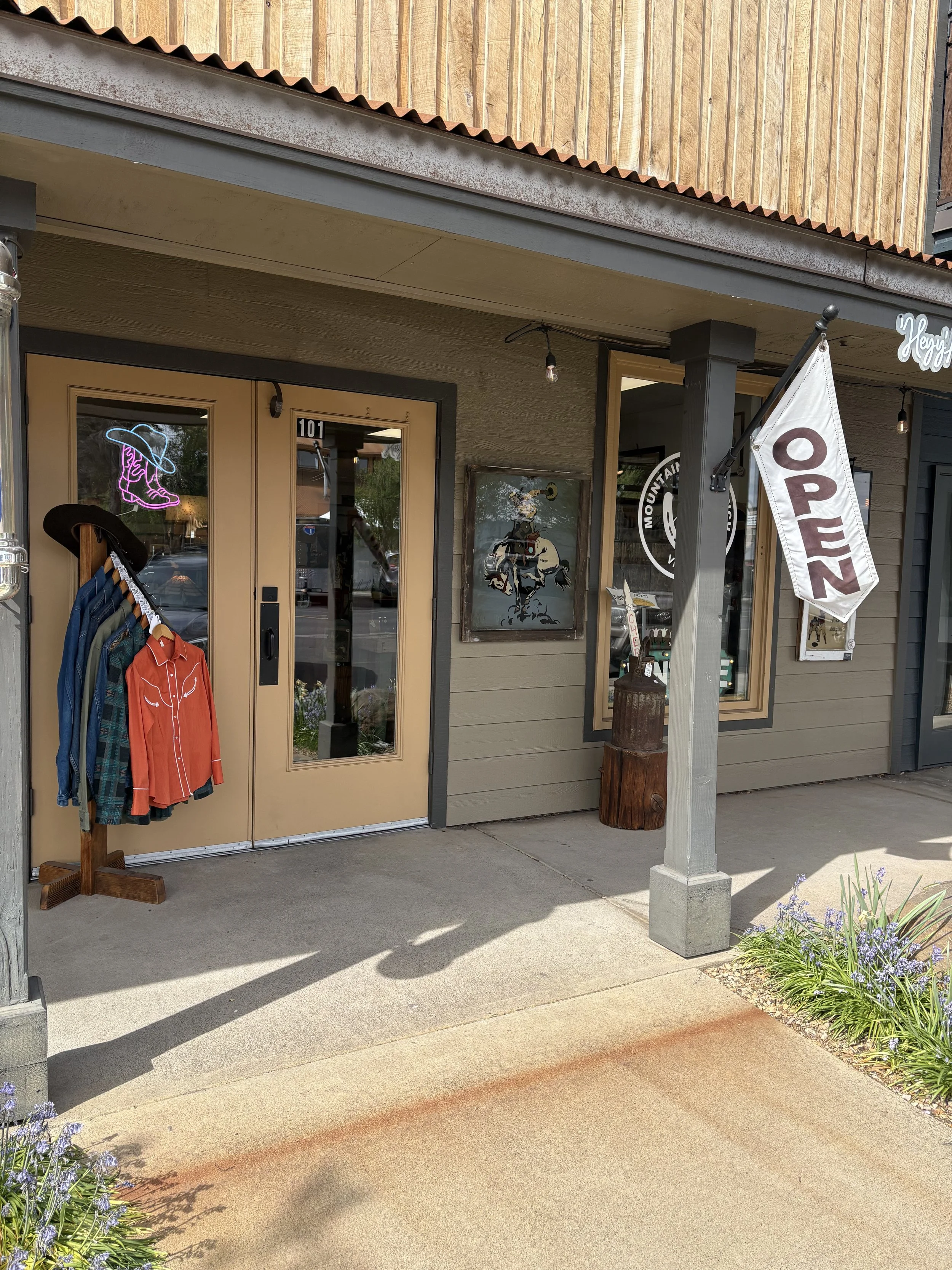 Outside view of a retail store with an "Open" sign on a flag, a clothing rack with jackets and a hat, and artwork on the wall near the entrance.