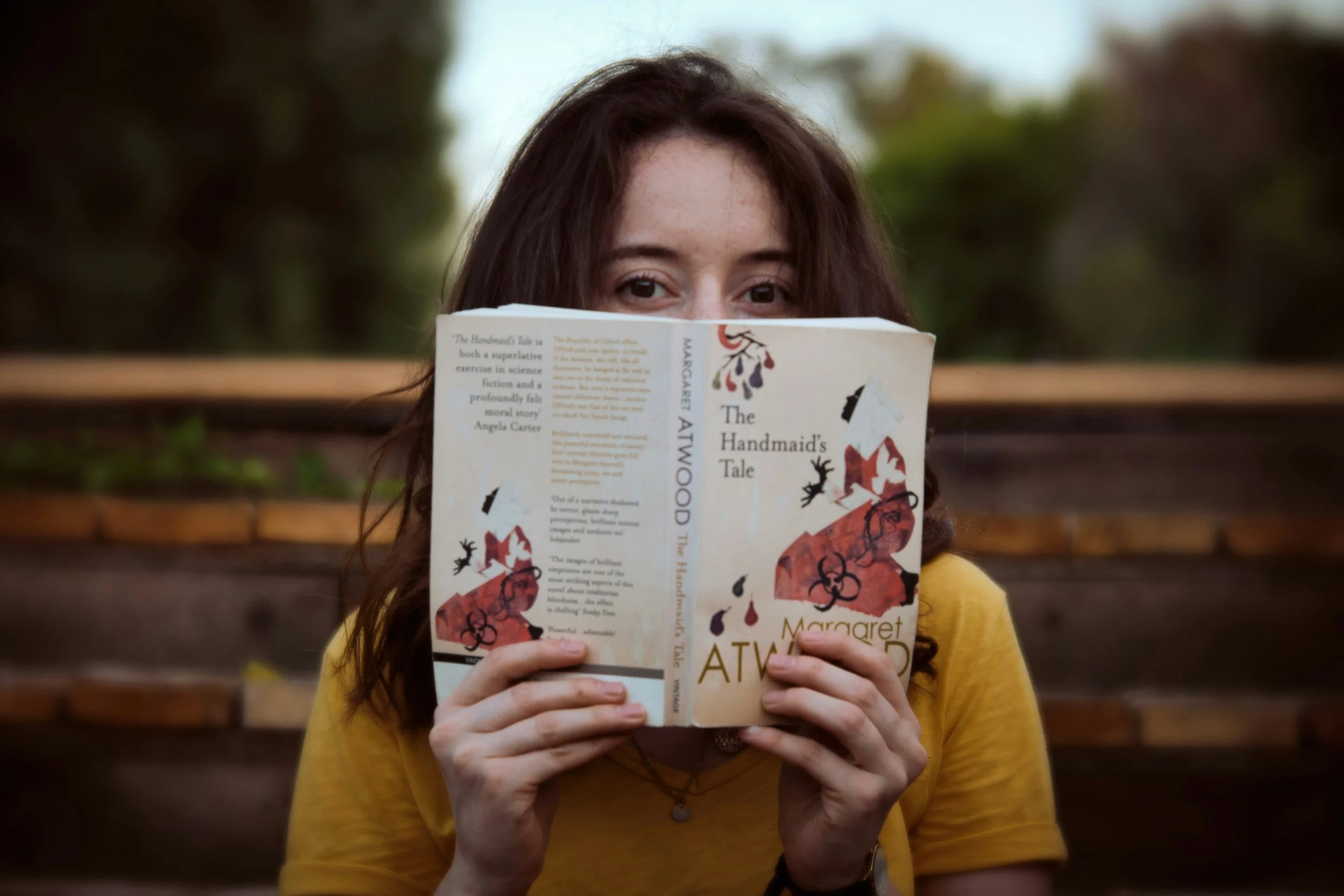 A young woman cheekily peeks over a copy of "The Handmaid's Tale" by Margaret Atwood.