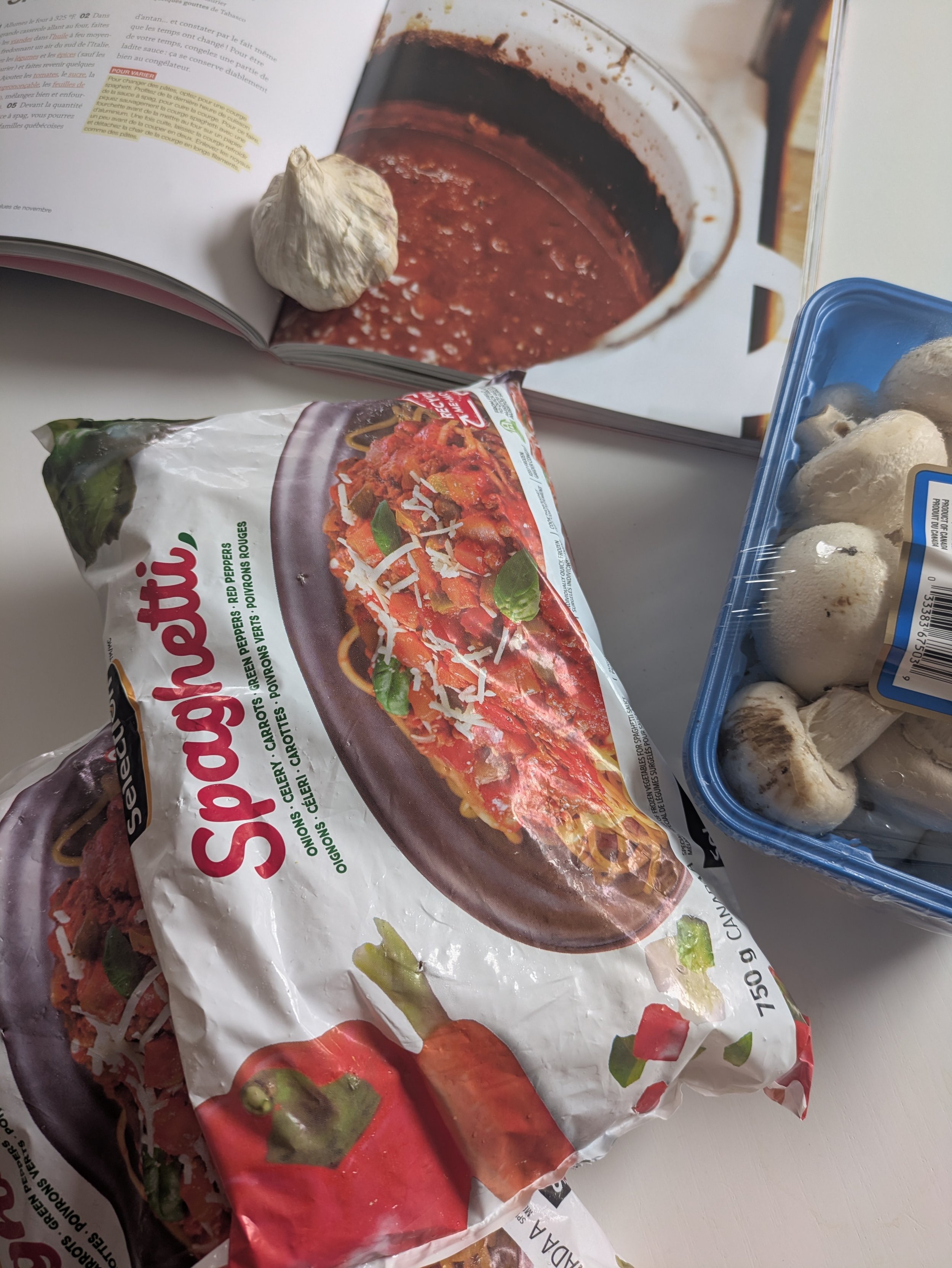 Des légumes congelés déjà coupés, des champignons, de l'ail et un livre de recettes pour faire du spaghetti