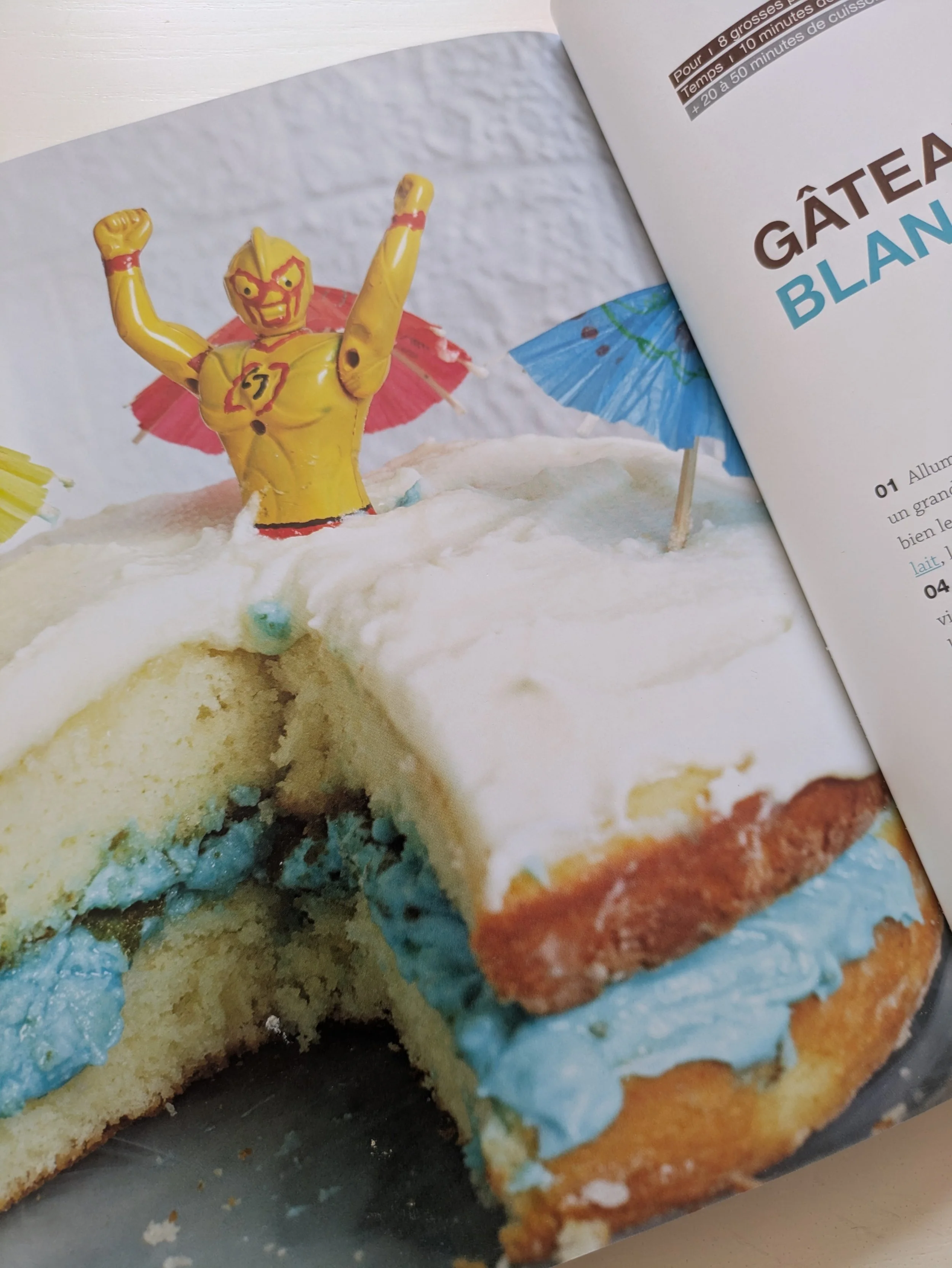 La photo d'une des pages du livre de la Croûte Cassée qui met en vedette un gâteau blanc avec un intérieur de glaçage bleu et une figurine de super héro planté dedans