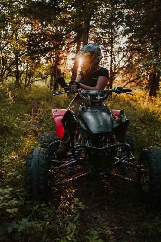 A person wearing a helmet sits on a black and red ATV in a forest during sunset.