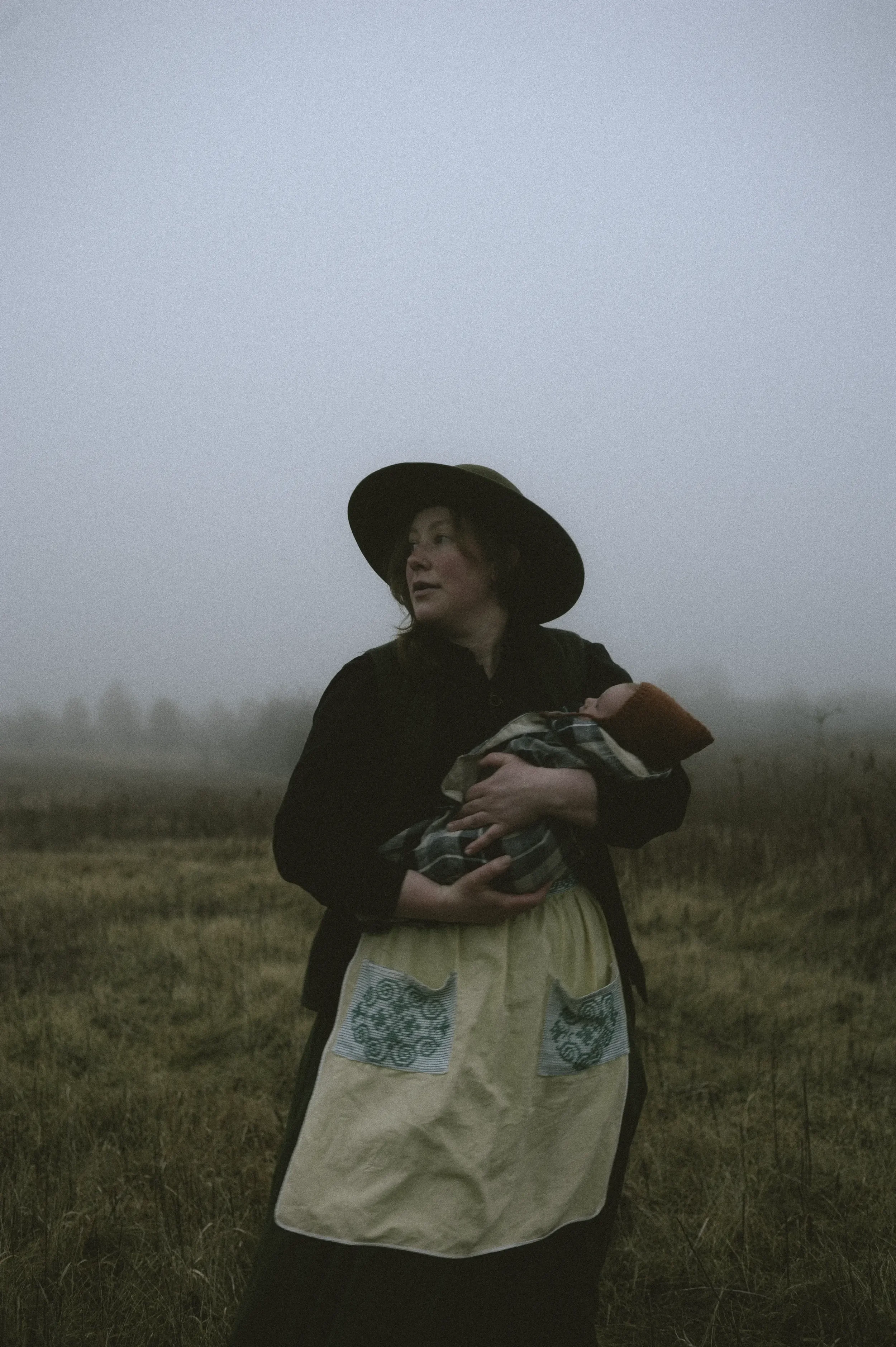 A woman in a wide-brimmed hat holding a baby wrapped in a blanket, standing in a foggy field.