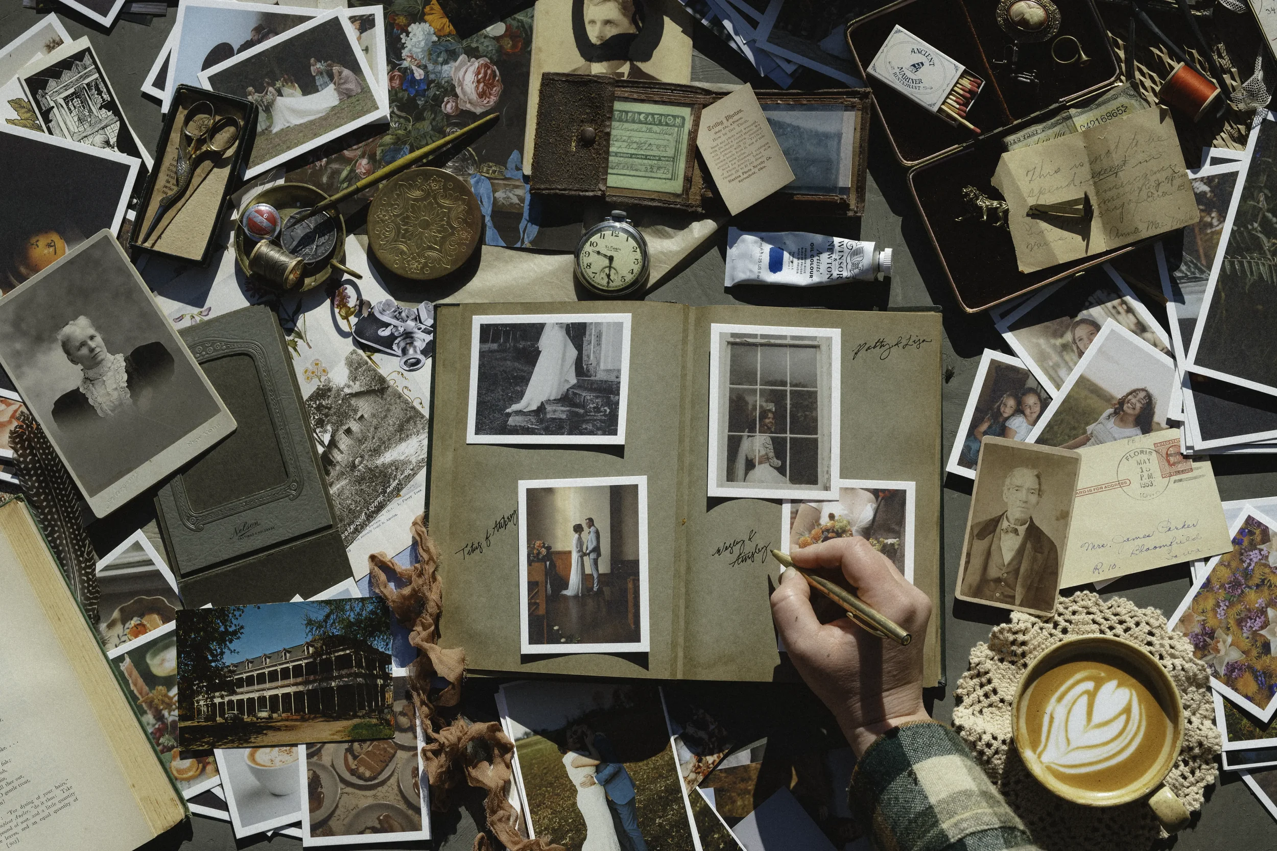 A person writes in a photo album surrounded by vintage photographs, postcards, and memorabilia on a table, along with a cup of coffee with latte art, a tube of cream, and various small vintage objects.