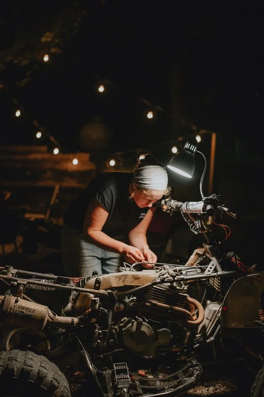A woman working on a motorcycle at night under a lamp, with string lights hanging in the background.