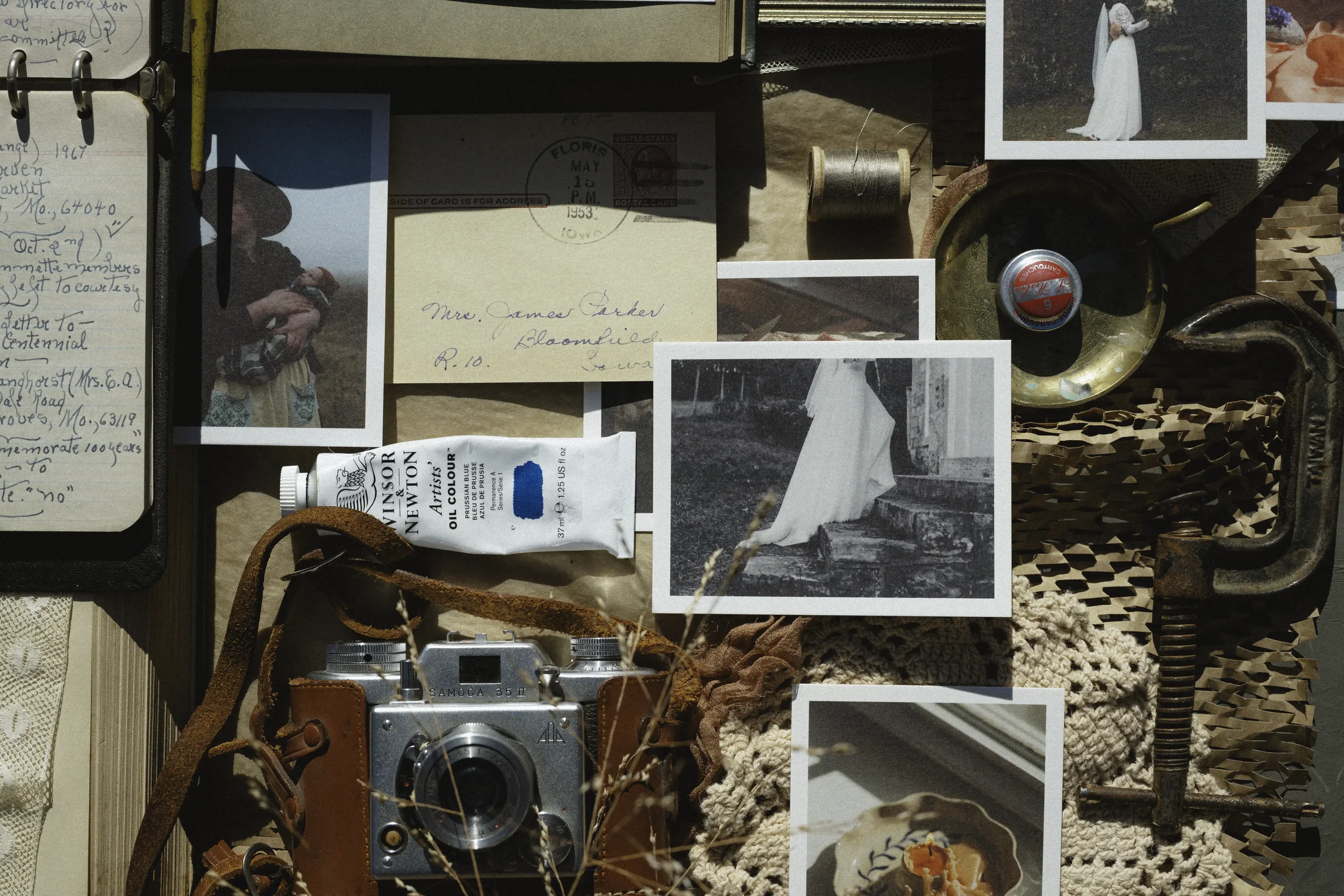 A collection of vintage photography and memorabilia including black and white photographs of a woman in a white dress, a woman in a hat holding a camera, and a scene with stairs, along with handwritten notes, a Kodak camera, a tube of oil paint, a spool of thread, a small metal tray, a rusty clamp, and a photograph of a bowl with an orange flower, all arranged on a textured surface.