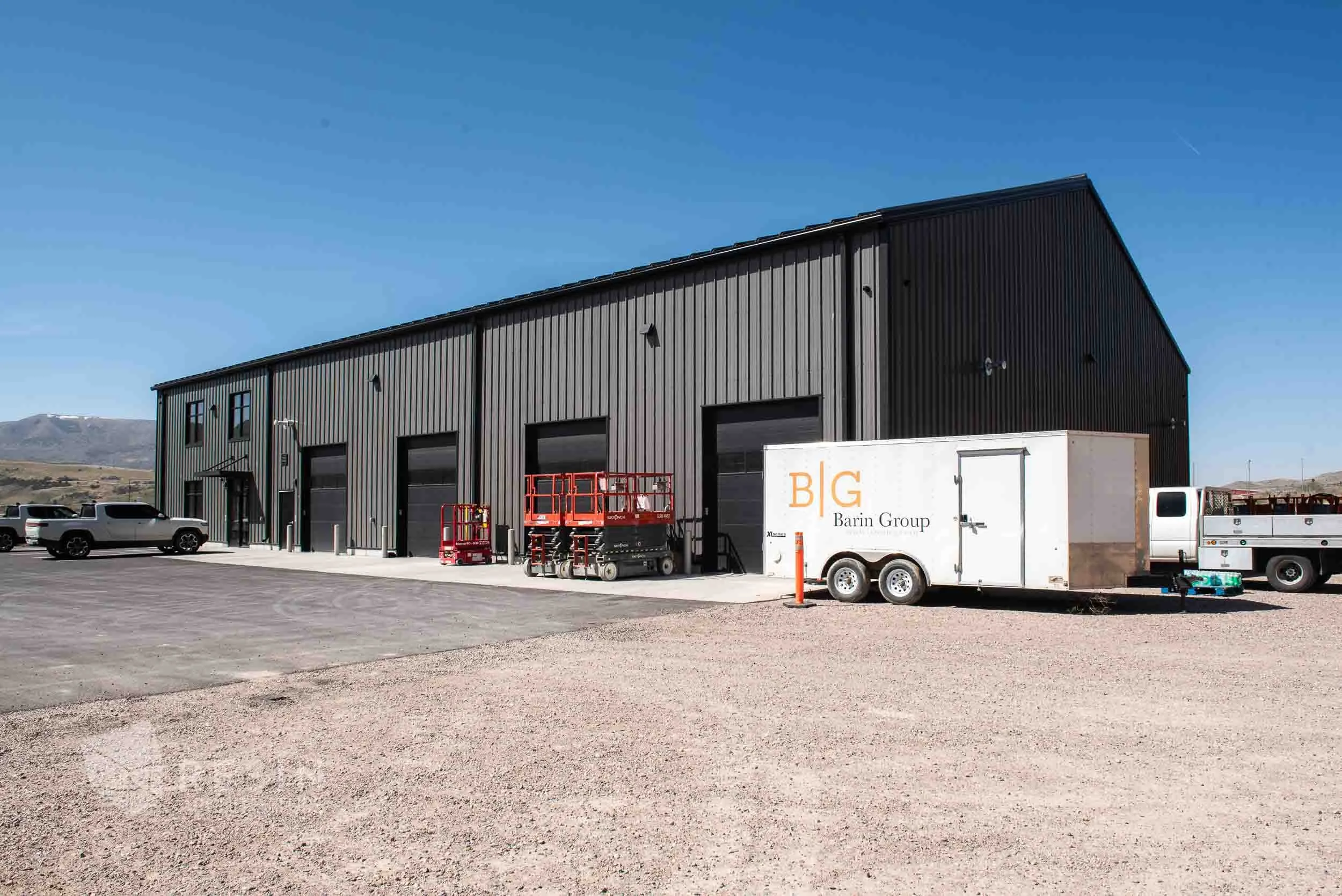 A large black industrial building with four garage doors, surrounded by vehicles including a white truck with a trailer, and red equipment. The setting is outdoors on a sunny day with mountains in the background.