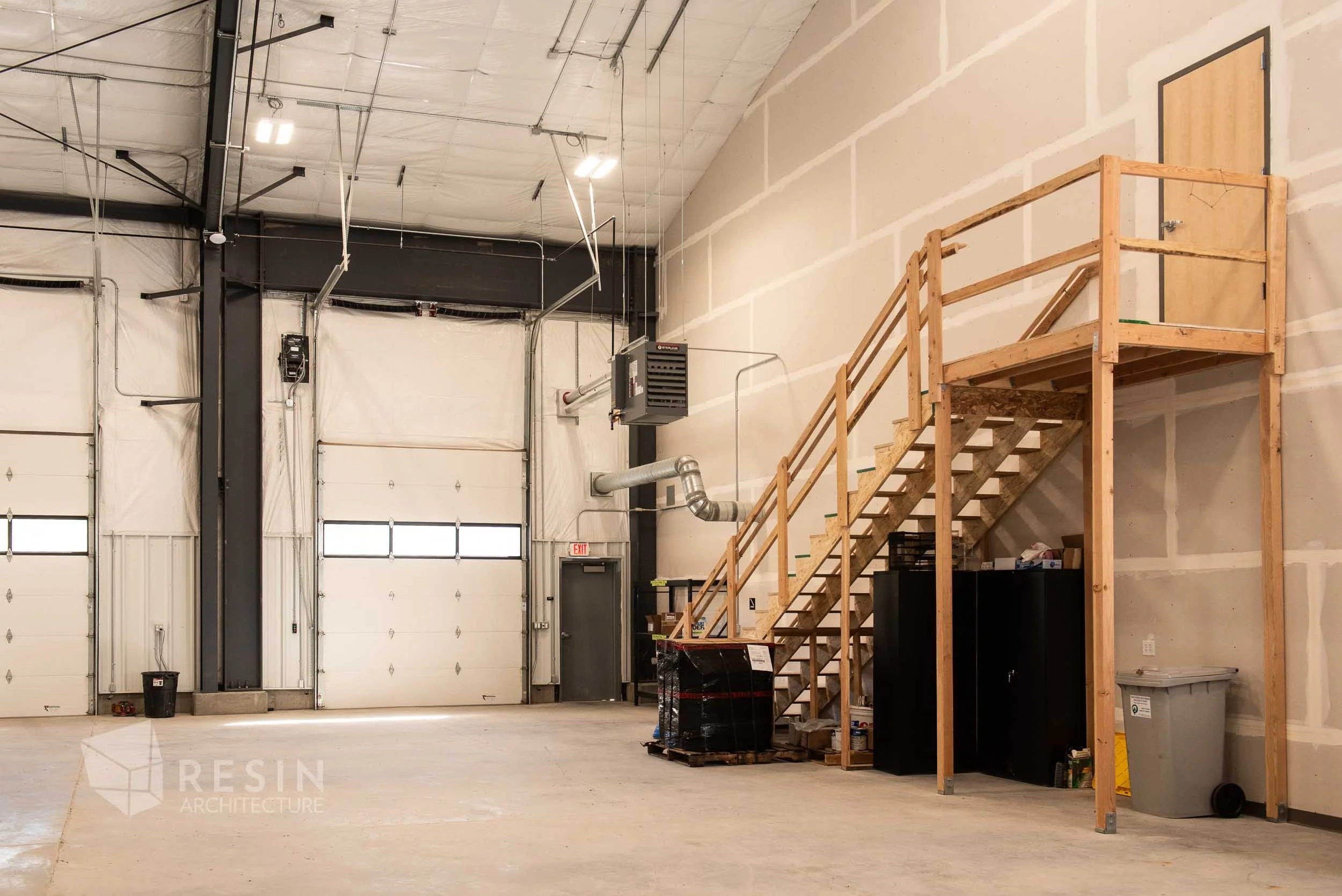 Inside a warehouse or industrial space with a large open floor, high ceiling, and white garage doors. There is a wooden staircase leading to a small upper platform, and various storage cabinets and containers are visible under the stairs. Exposed duc