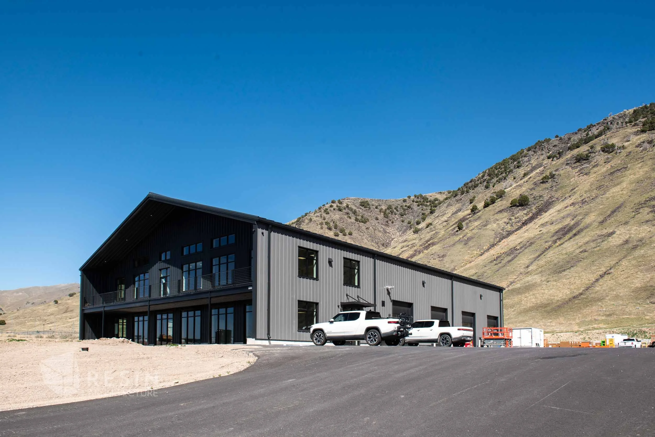 Modern gray industrial building in a rural area with hills in the background, parked white trucks, and clear blue sky.