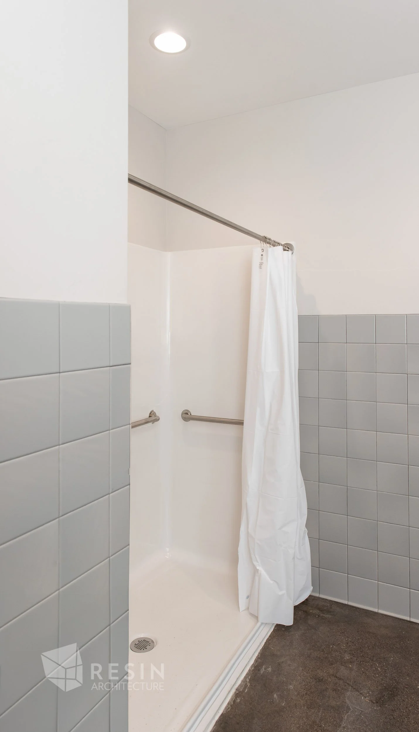 A small, accessible shower with white walls, gray tiled walls, a stainless steel shower curtain rod, a white shower curtain partially closed, and a dark floor.