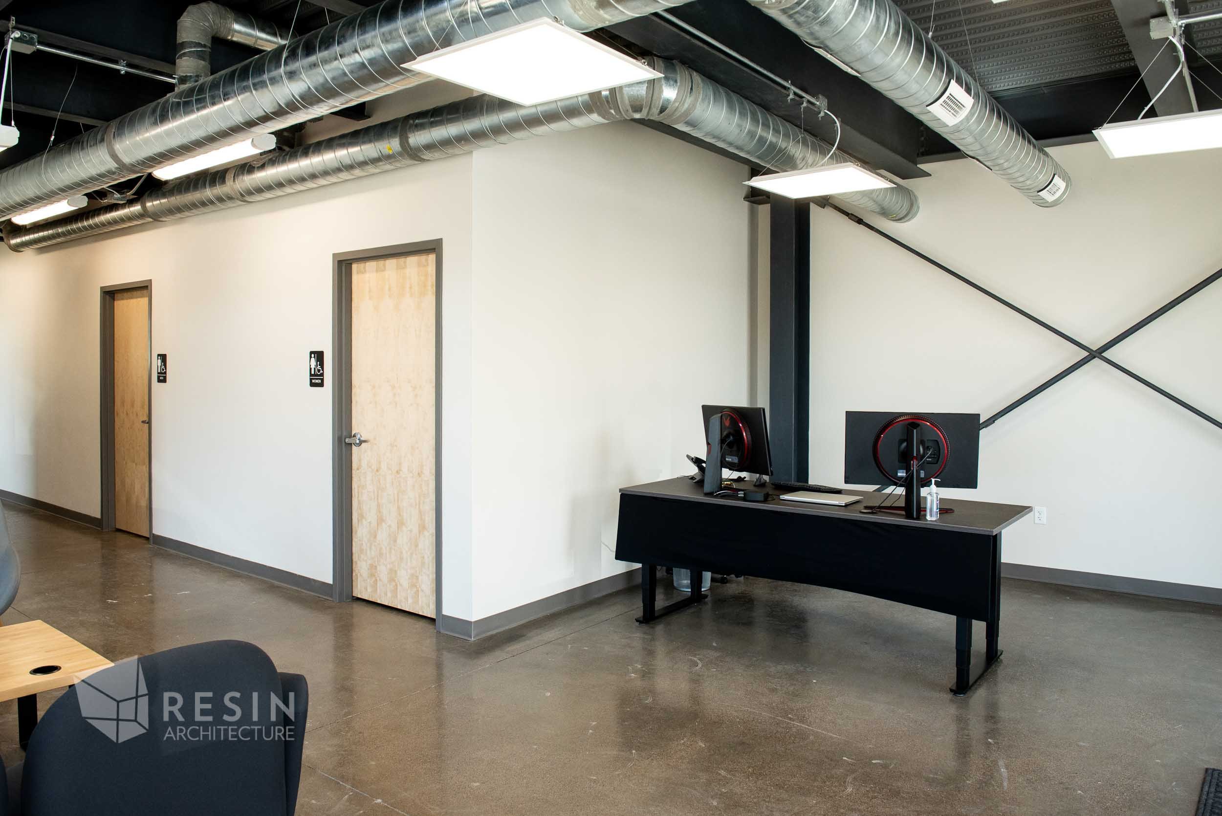 Modern office with two closed wooden doors, wall signs indicating restrooms, a black desk with computer monitors, and visible industrial ceiling ducts and lighting.