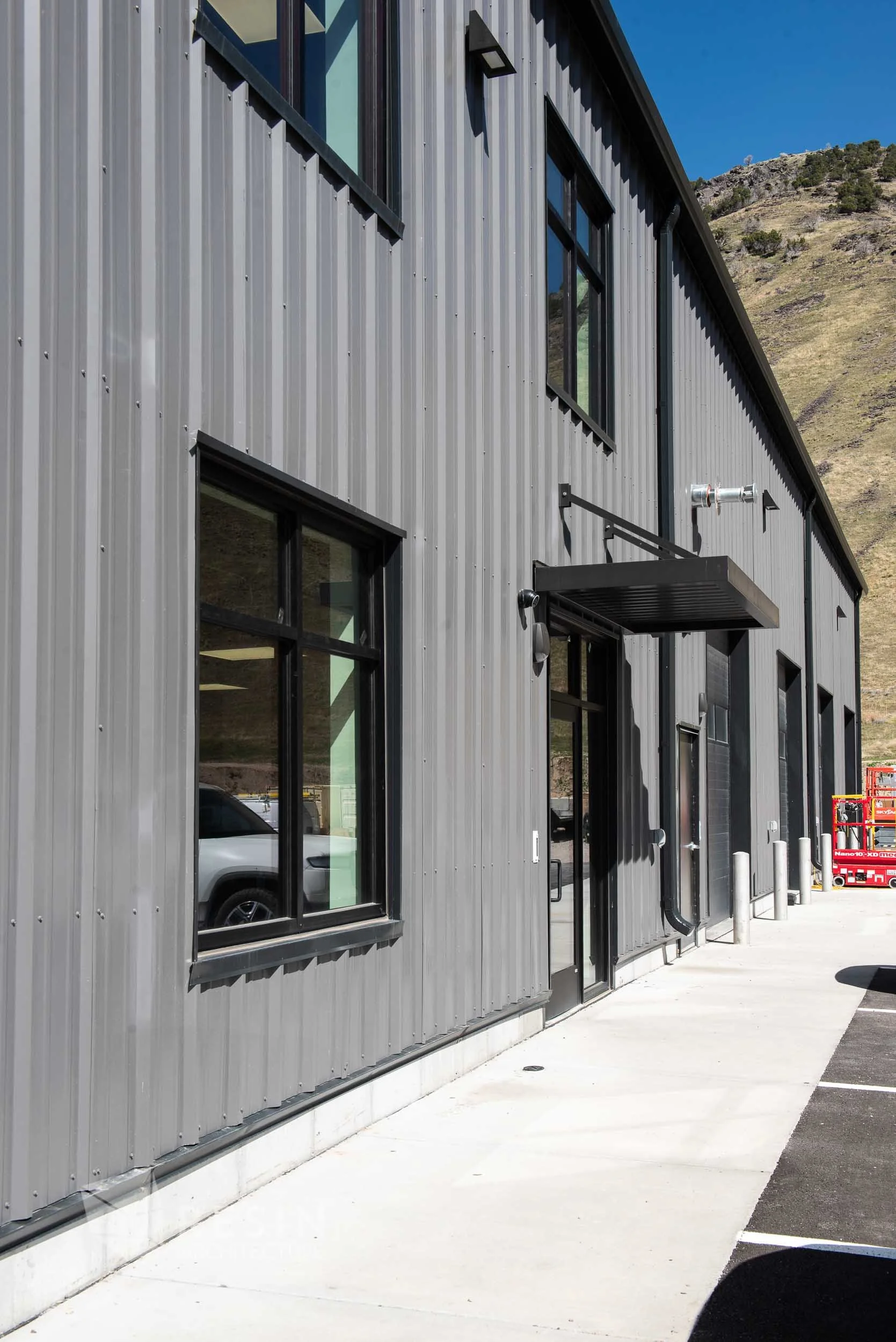 Modern metal building with large windows, a black door with a canopy, and a parking lot with a red lift in the background, mountains in the distance.